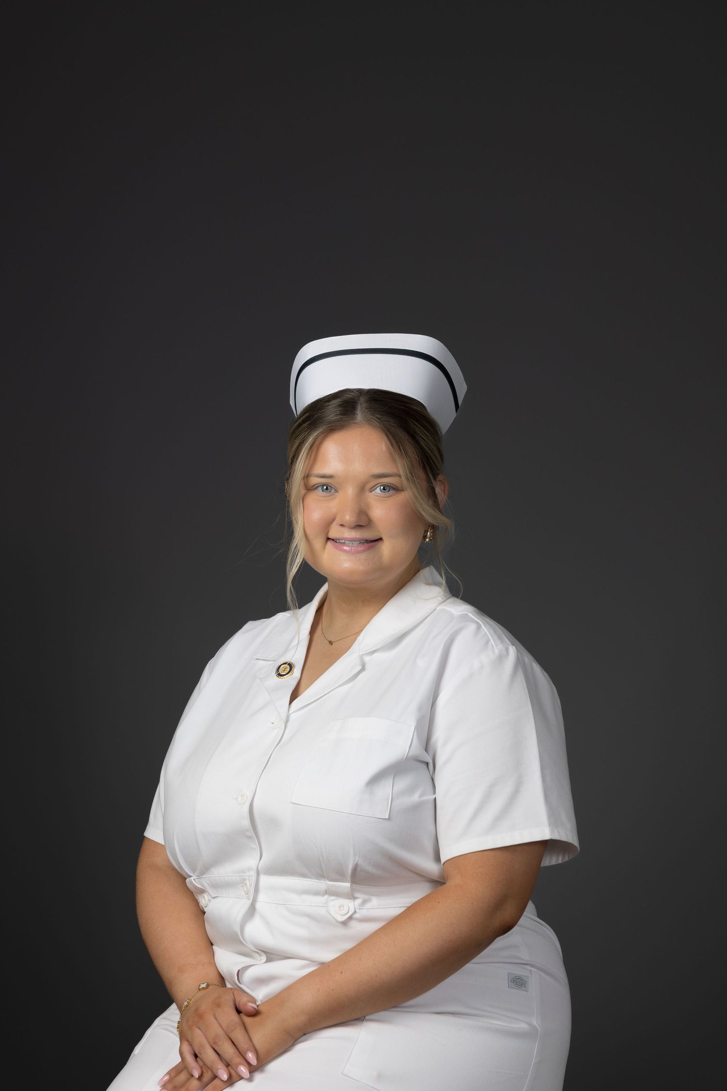 Smiling nurse in white uniform and traditional cap poses for a professional portrait against a gray background.