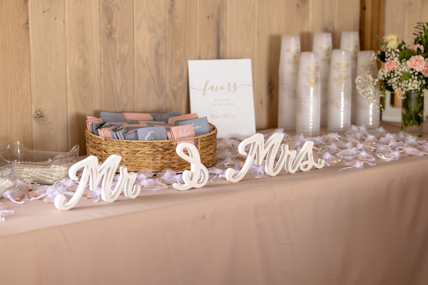 Wedding reception table with Mr. & Mrs. signs, wicker basket of favors, candles, and floral arrangement on blush linen.