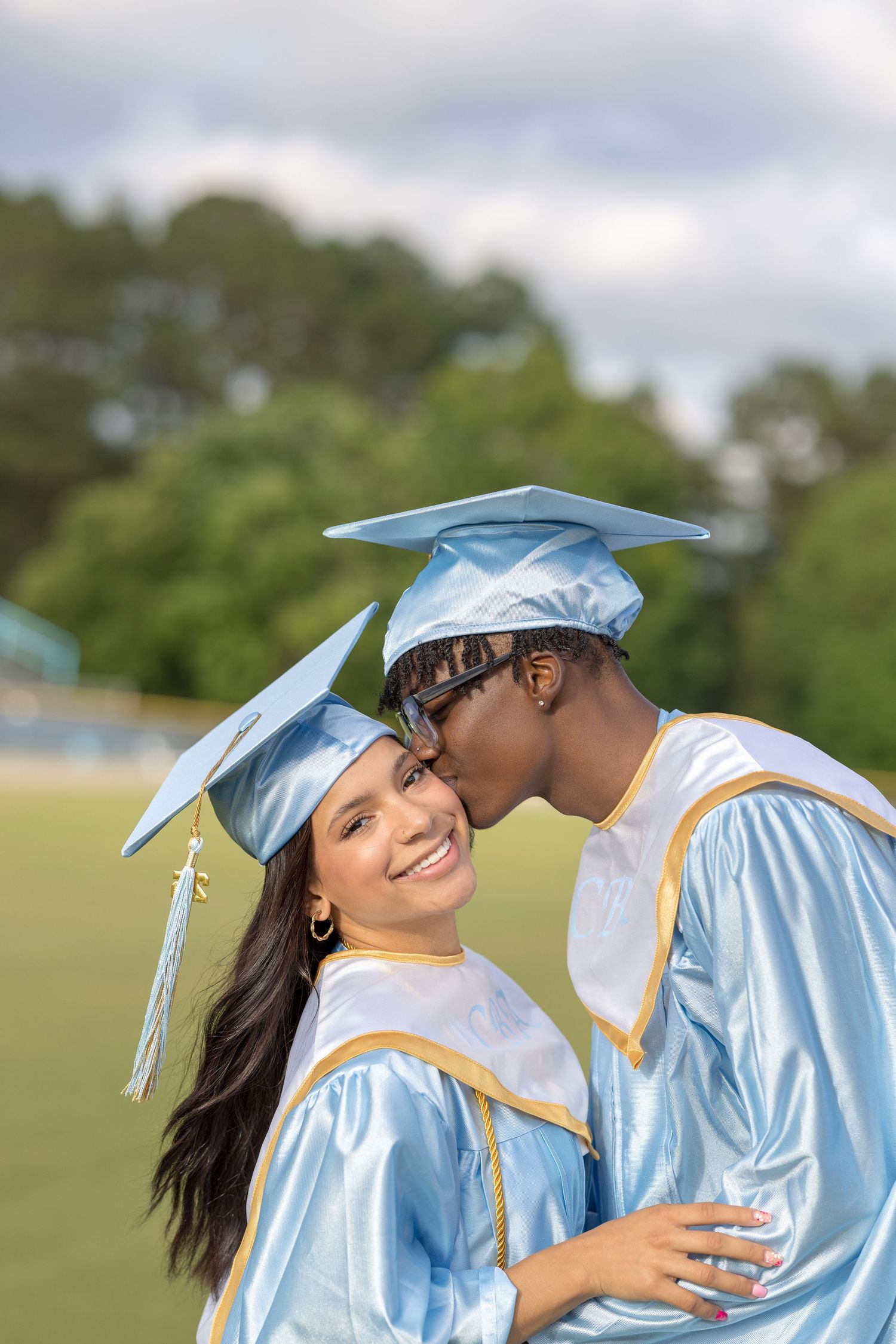 Two graduates in light blue caps and gowns share an intimate moment on a sunny day with green trees in background.