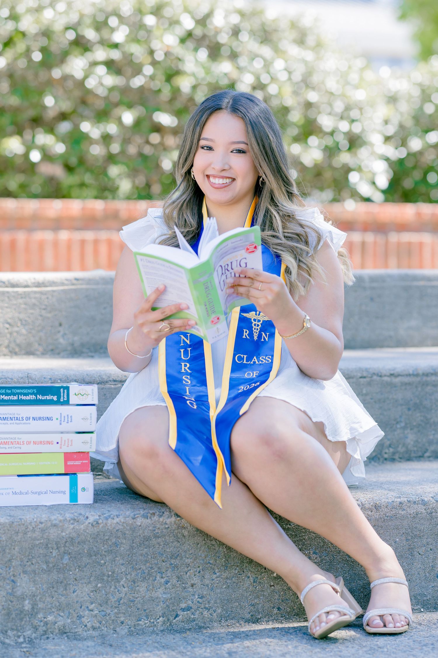 Smiling nursing graduate sits on steps holding a drug guide book, wearing blue RN sash and white dress with stacked textbooks beside her.