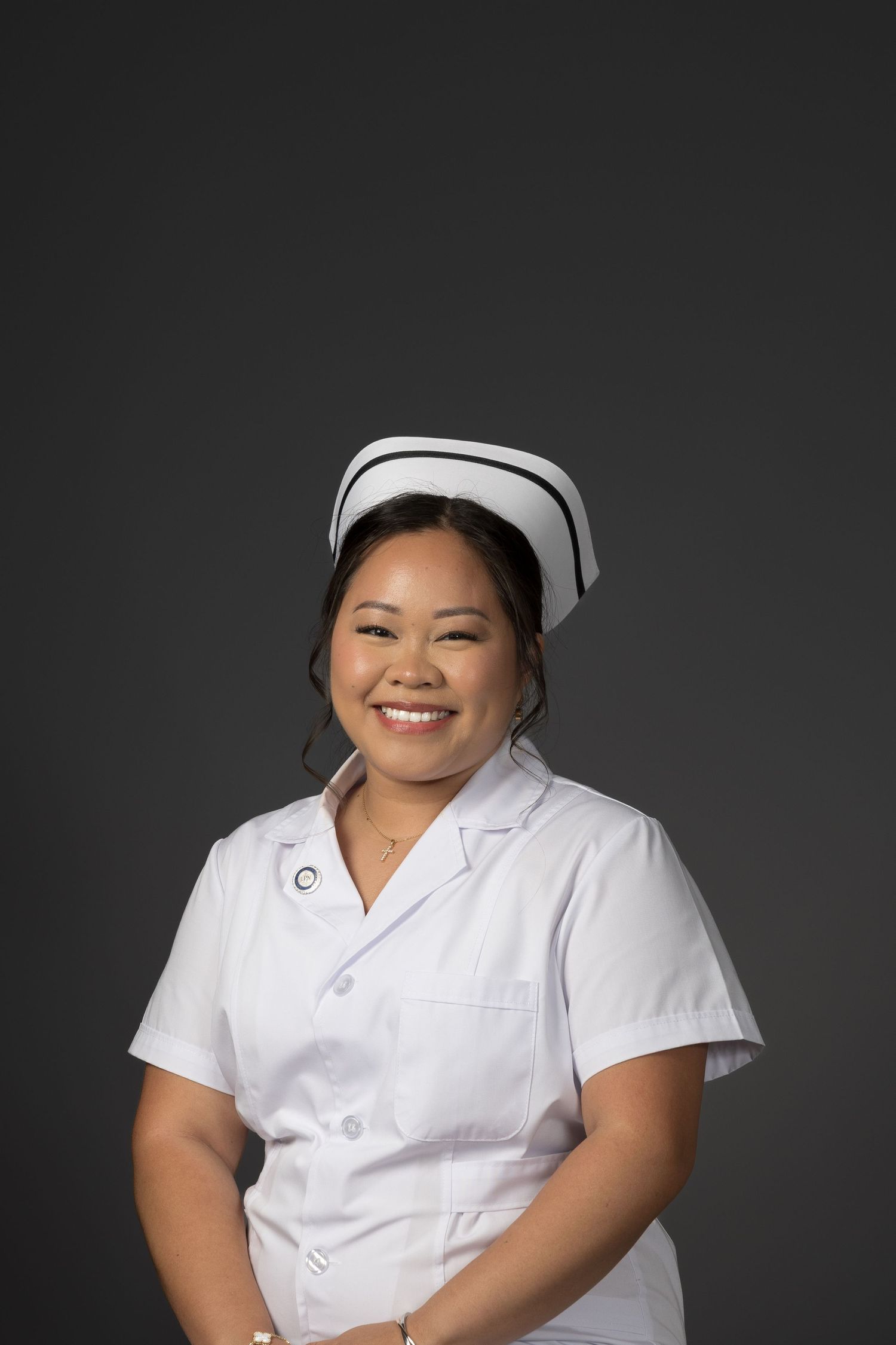 Smiling Asian female nurse in white uniform and cap posing against gray background.
