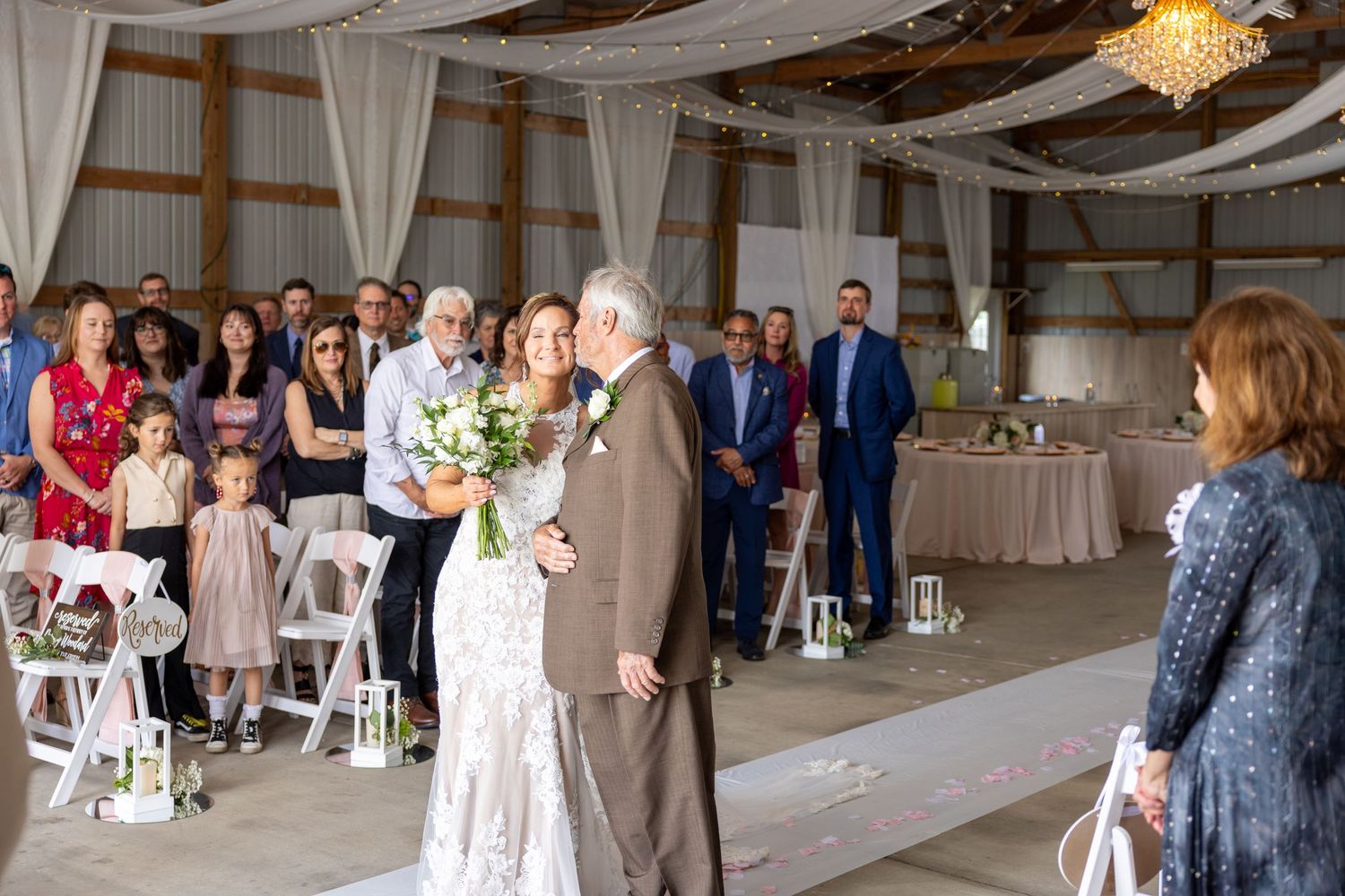 Bride walks down the aisle with her father in a rustic barn wedding venue surrounded by standing guests.