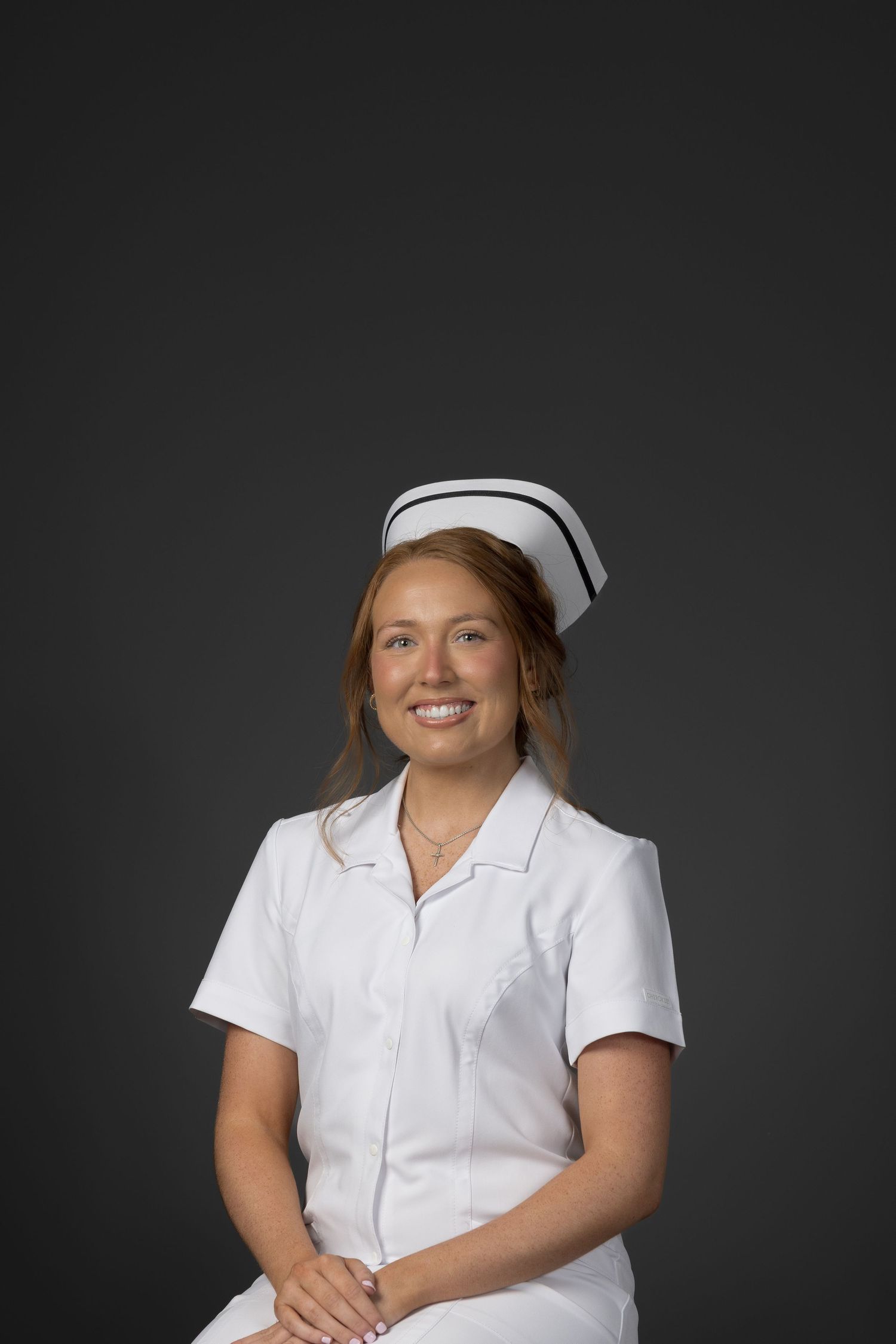 Smiling female nurse in white uniform and cap posing against gray background.