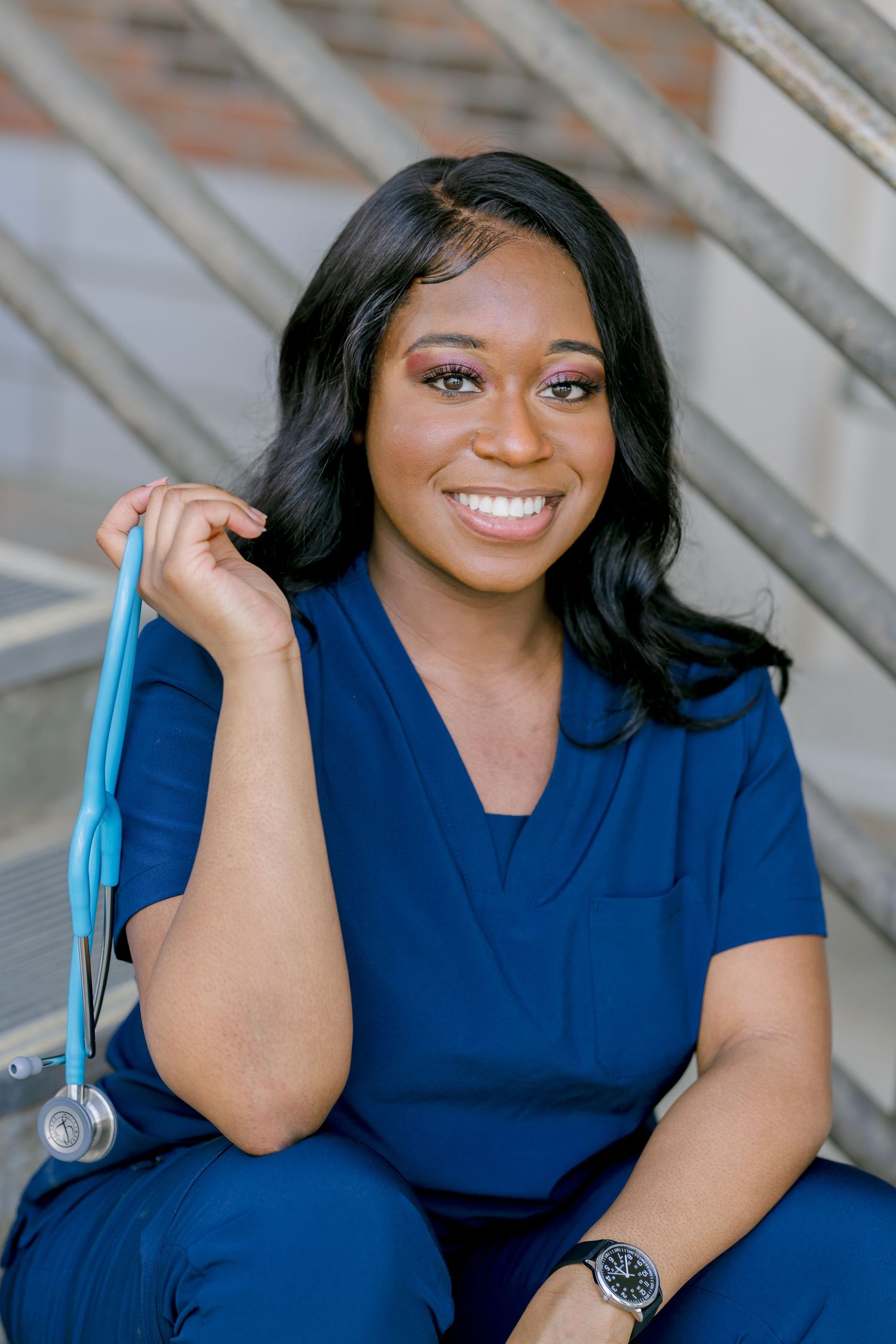 Healthcare professional in navy blue scrubs smiling while sitting on concrete steps outdoors.