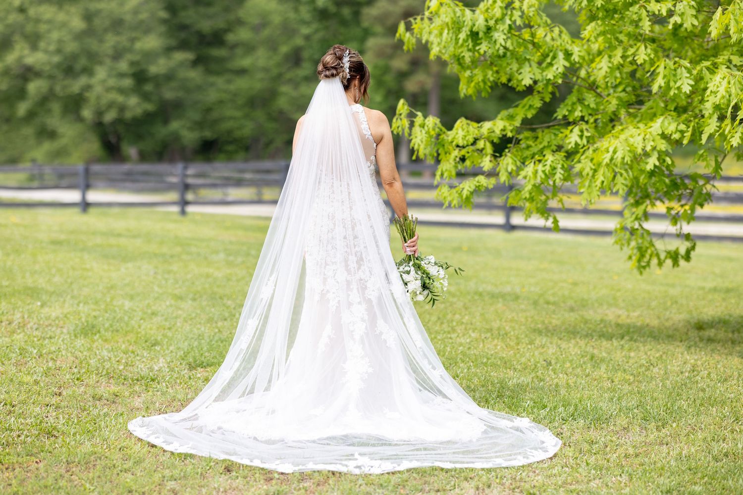 Bride in white gown with long veil holding bouquet, standing on green lawn near trees.