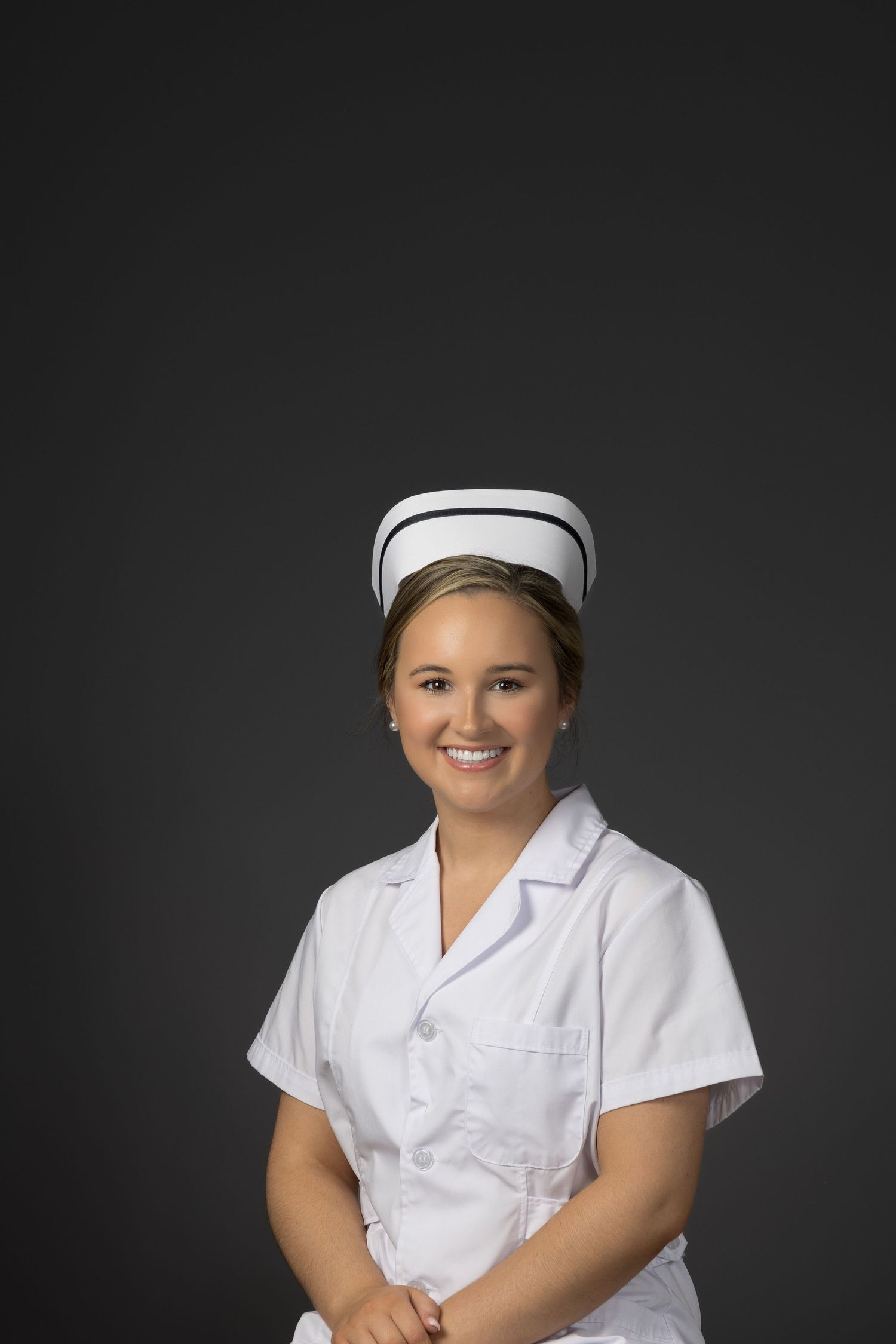 Smiling young female nurse in white uniform and traditional cap posing against dark gray background.