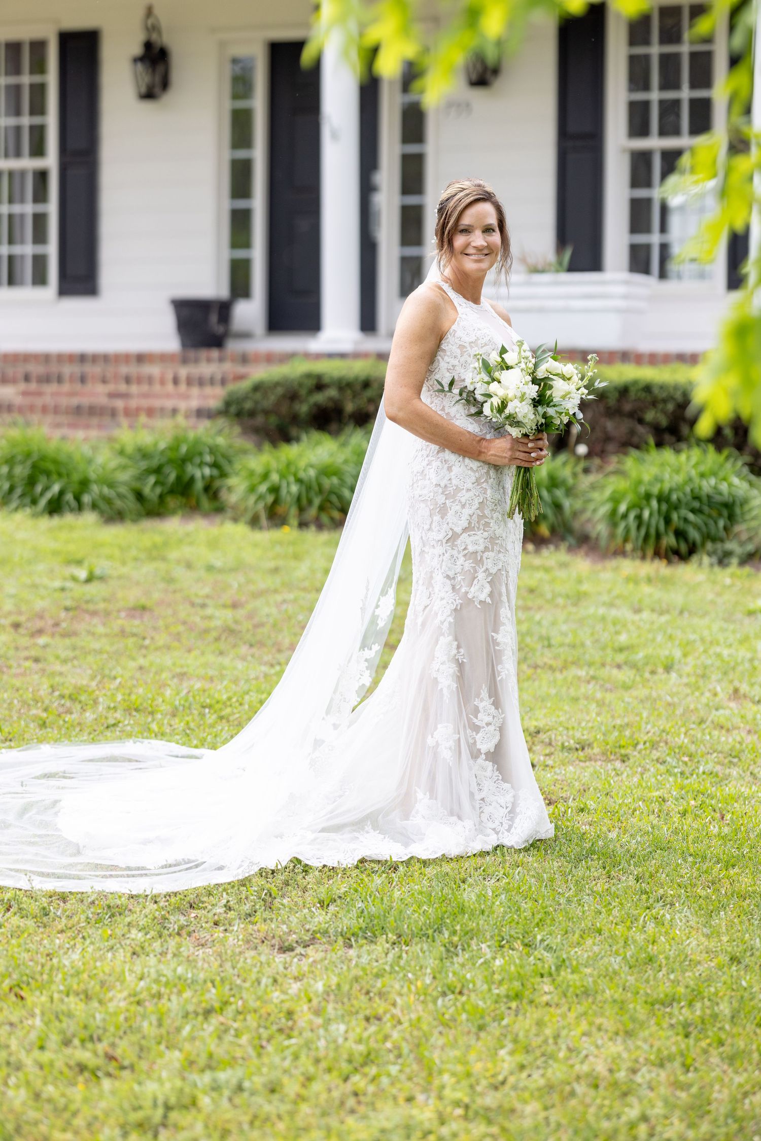 Bride in lace gown with veil holding white bouquet, standing on green lawn before white colonial home.