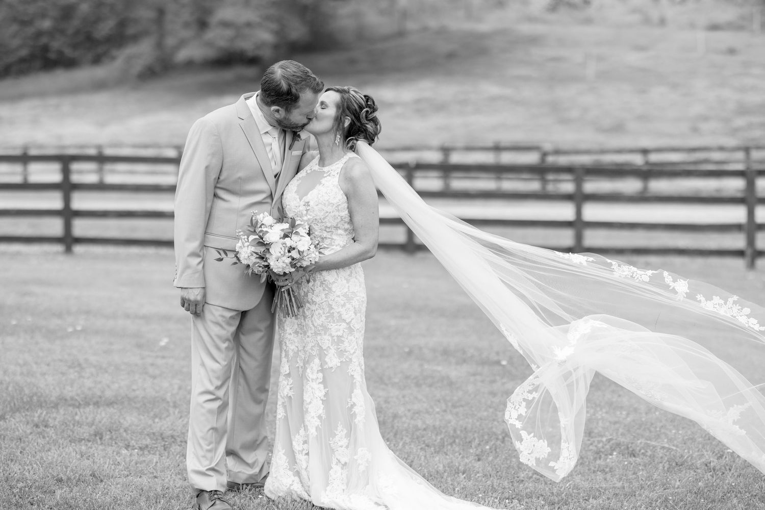 Black and white photo of a bride and groom kissing outdoors, her lace veil flowing in the wind near a rustic fence.