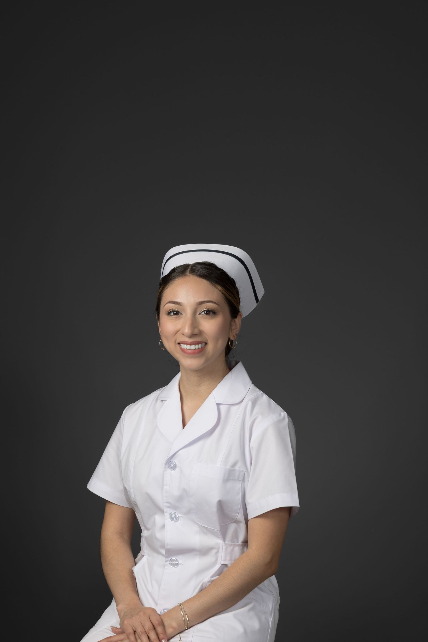 Young female nurse in white scrubs and cap smiling confidently against gray backdrop.