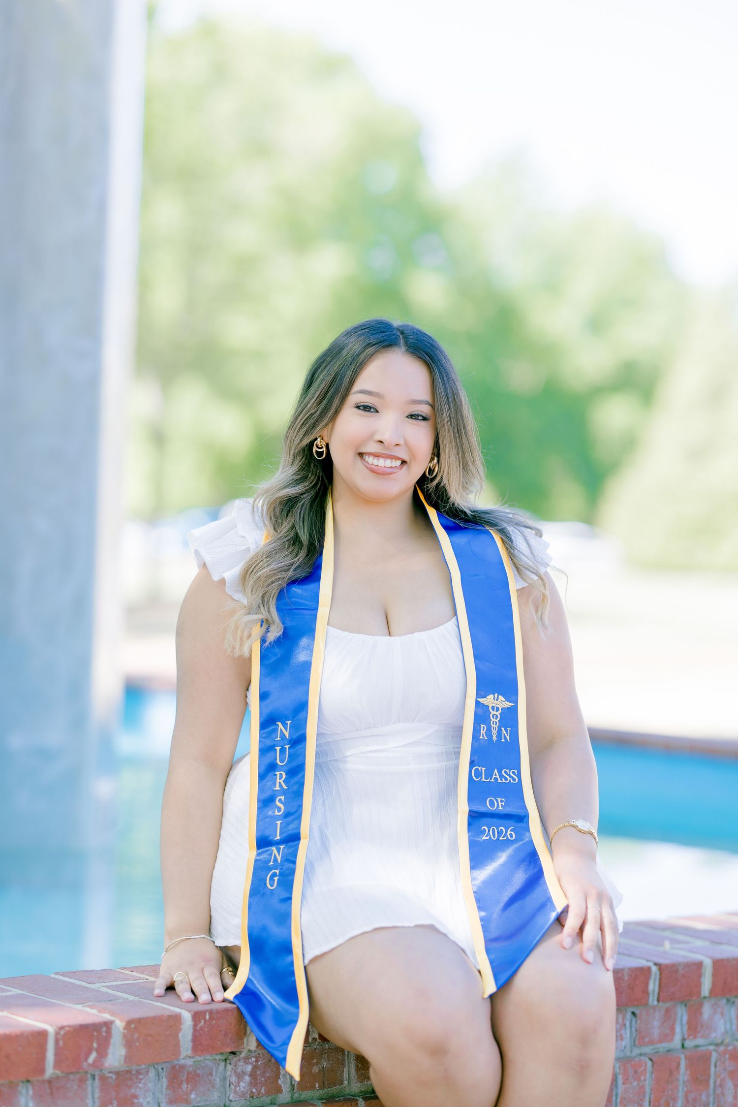 Smiling nursing graduate poses outdoors in white dress with blue and gold Class of 2026 nursing stole.