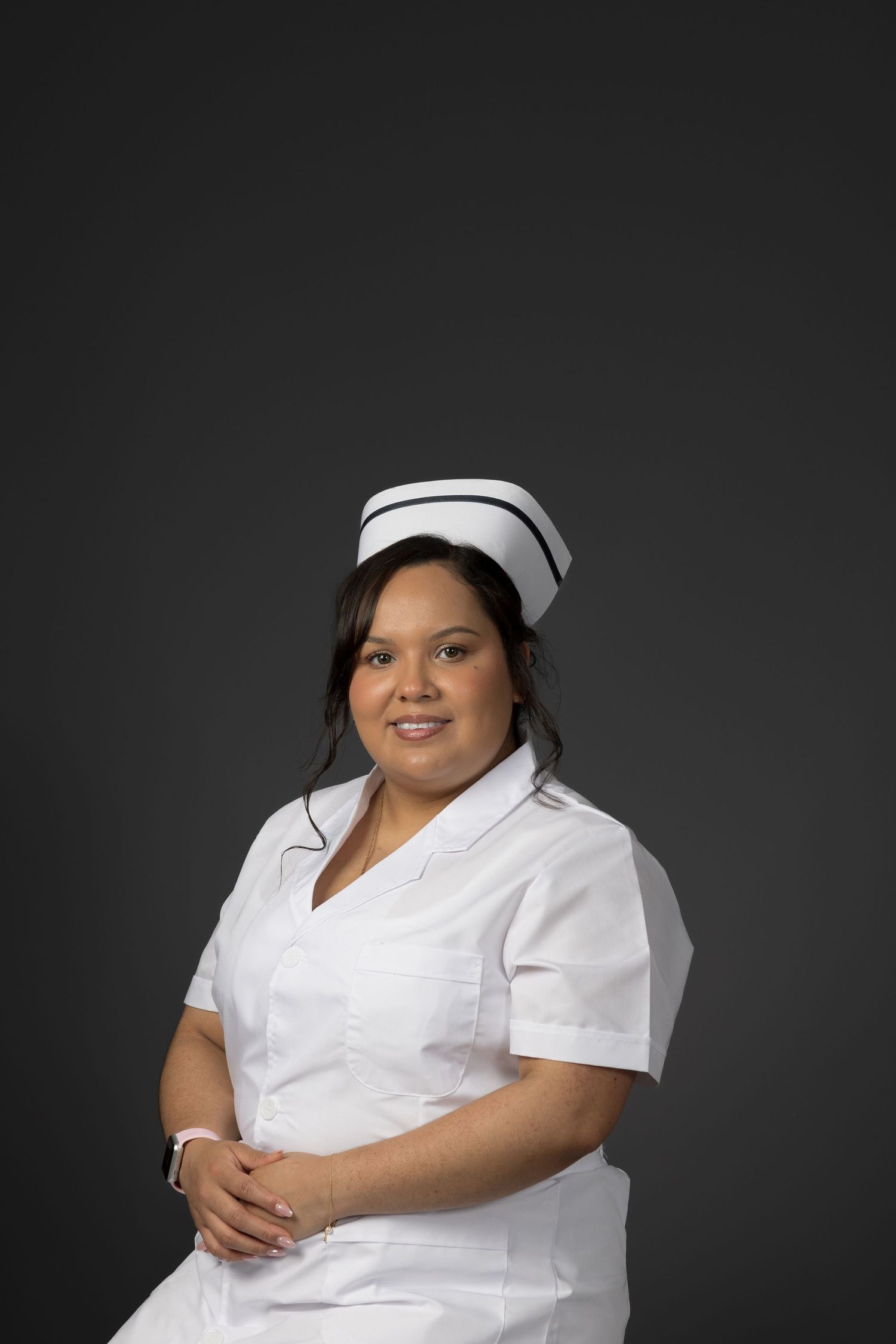 Smiling female nurse in white uniform and cap posing with hands clasped against dark gray backdrop.