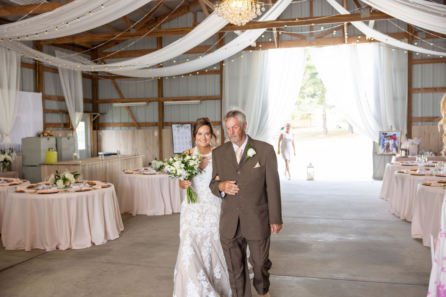 Bride and groom walking arm in arm through a decorated barn wedding venue with draped white fabric and floral centerpieces.