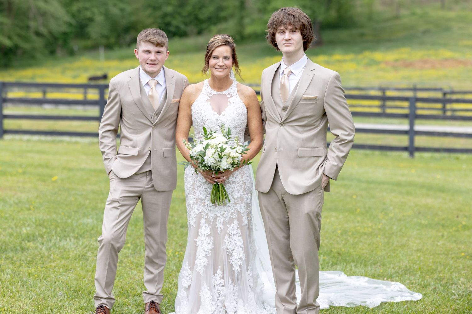 Bride in lace wedding gown holding bouquet stands with two groomsmen in tan suits on green grass.