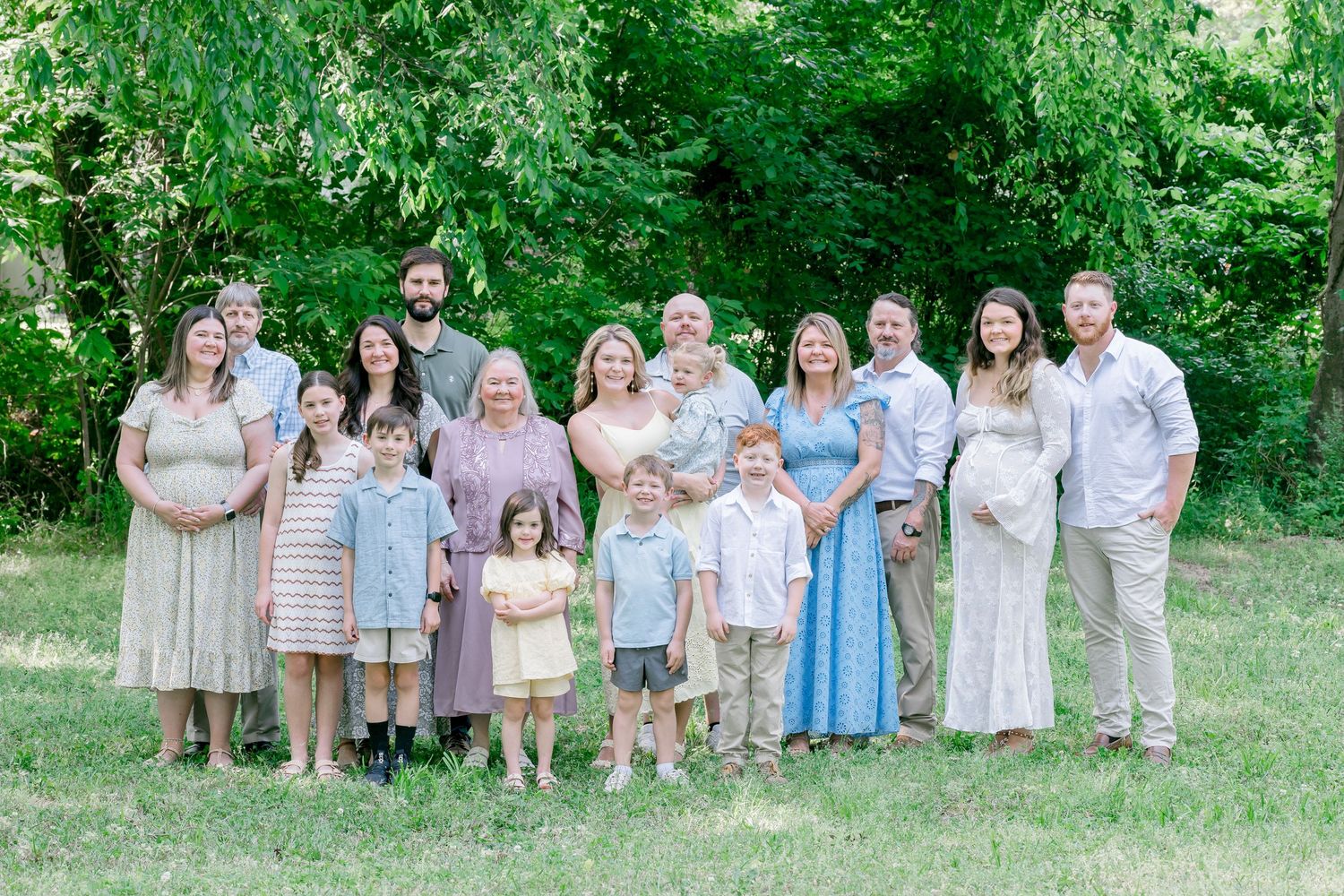 Large family group portrait outdoors on green grass with lush trees, featuring adults and children in coordinated pastel outfits.