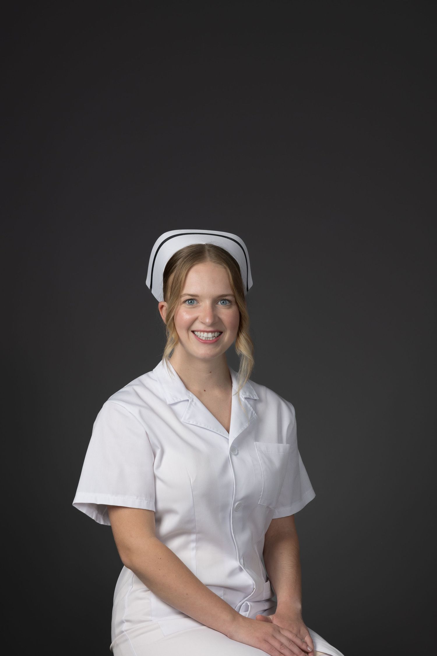 Young blonde nurse smiling in white uniform and cap against gray background.