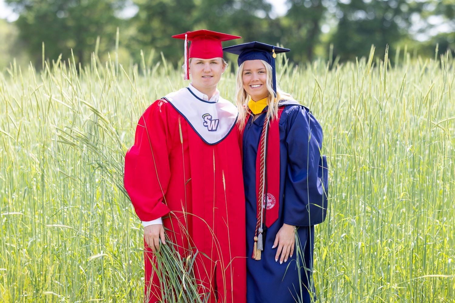 Two graduates in red and blue caps and gowns stand together in a sunny field of tall grass.