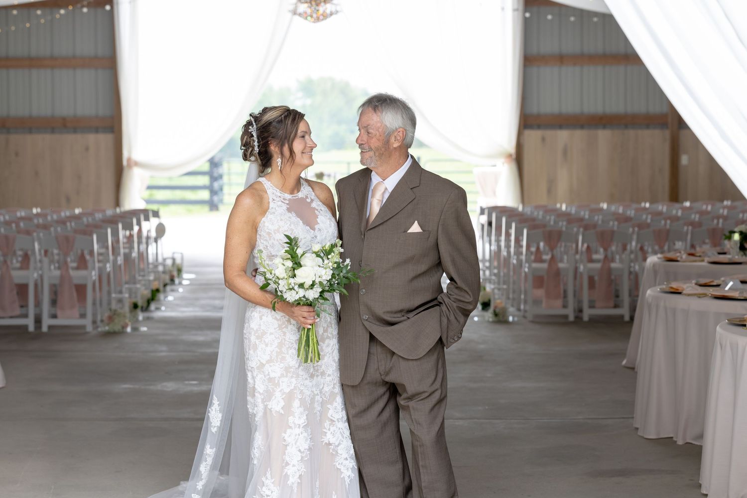 Bride and father share a tender moment in a rustic barn wedding venue with white draping and clear chairs.