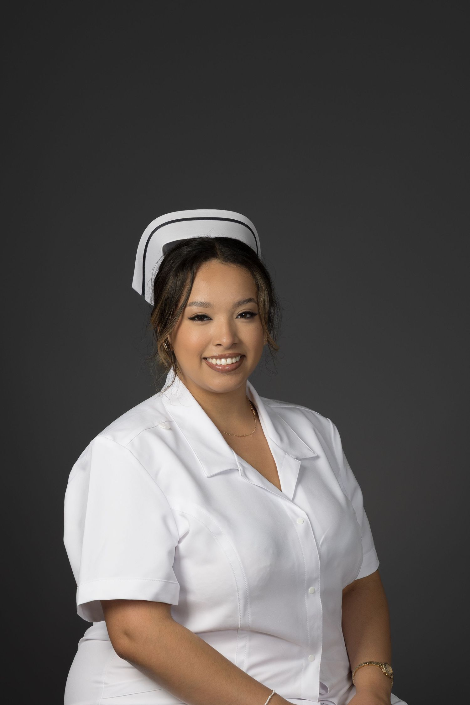 Smiling young woman in white nurse uniform and cap poses confidently against a gray studio background.