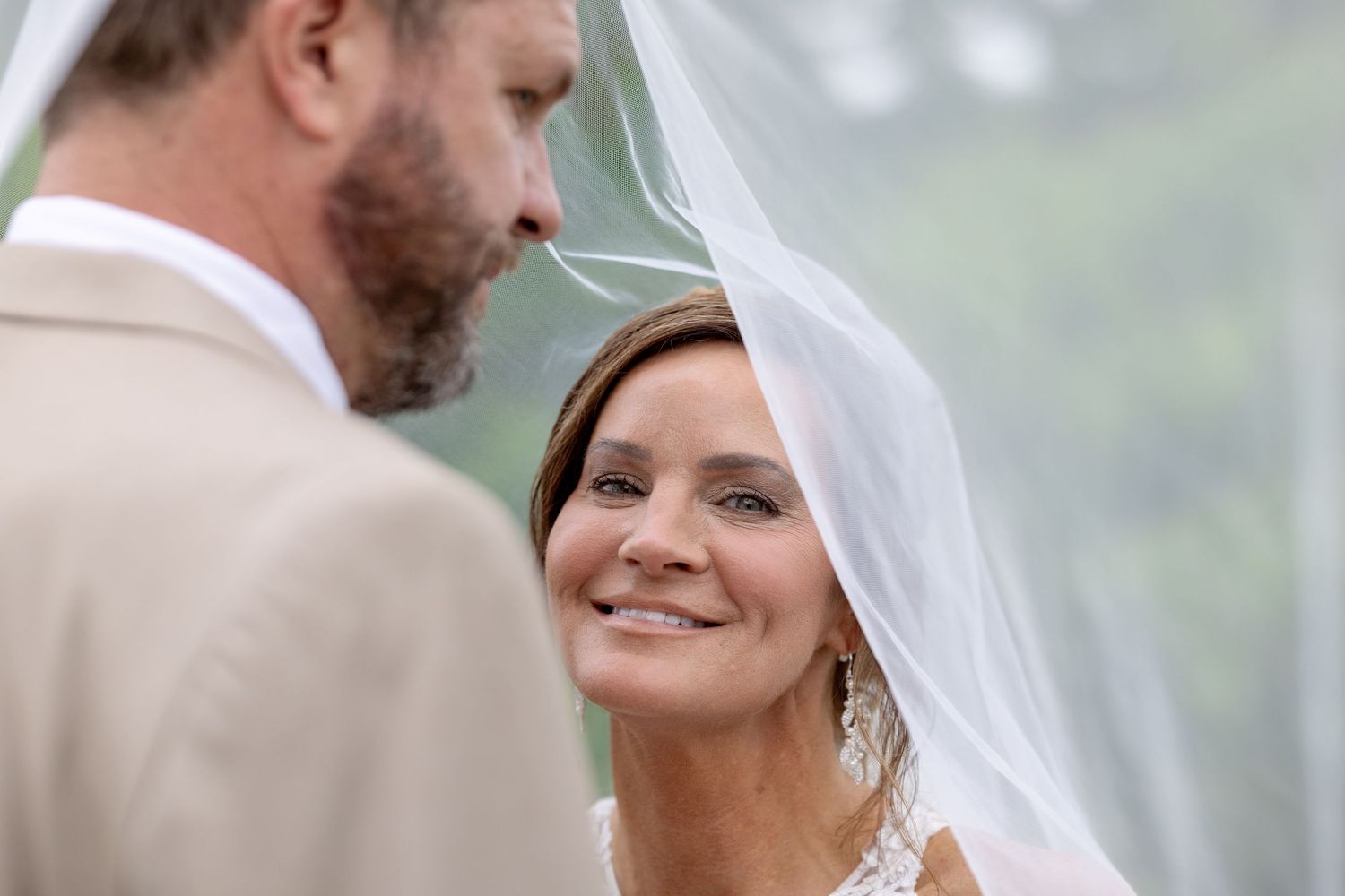 Bride in white veil smiling at groom in tan suit during outdoor wedding ceremony.
