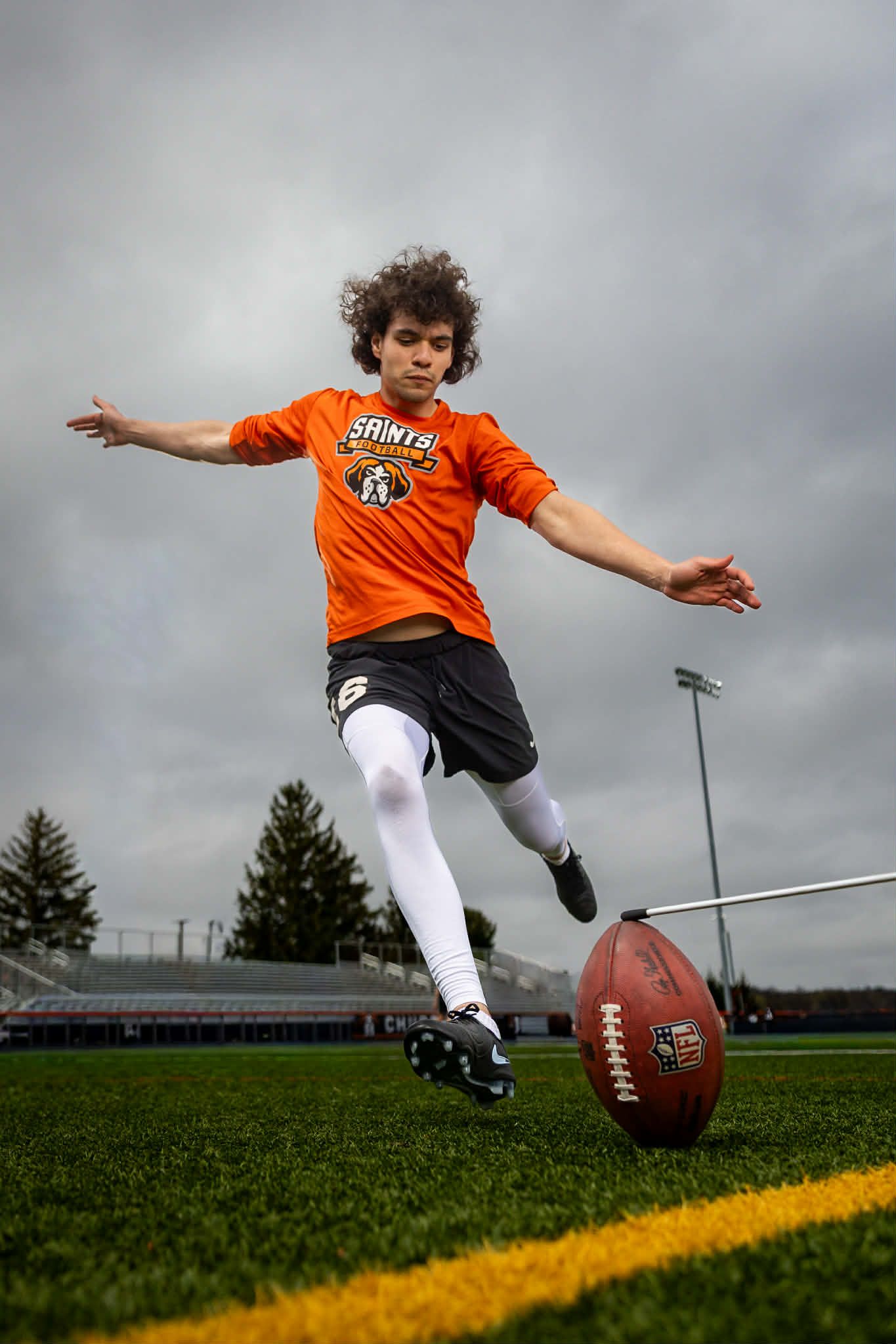 Young football player in orange Saints jersey kicking a football on a turf field under cloudy skies.