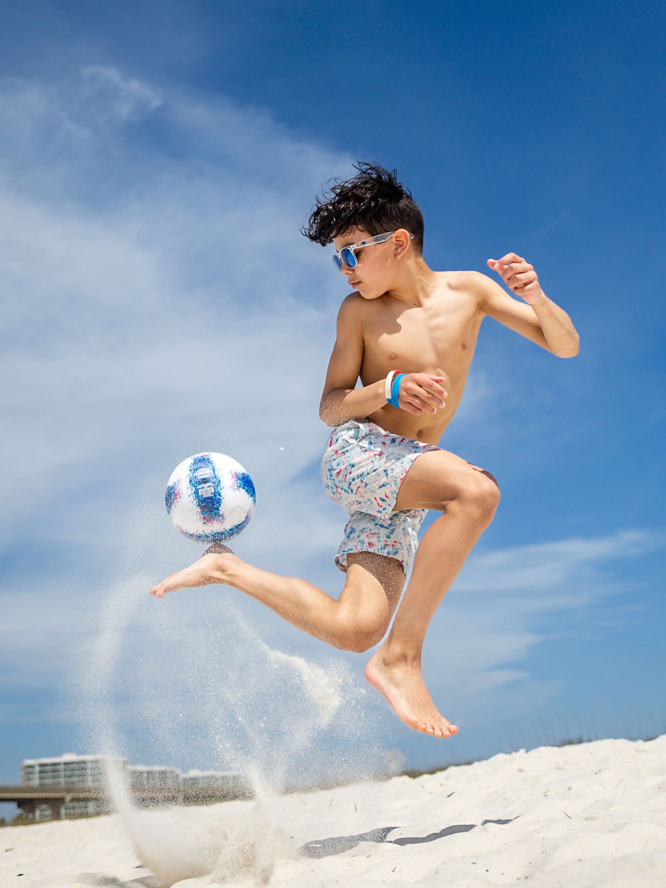 Boy in swim trunks and sunglasses jumps to kick a soccer ball on a sunny beach, sand flying around his feet.