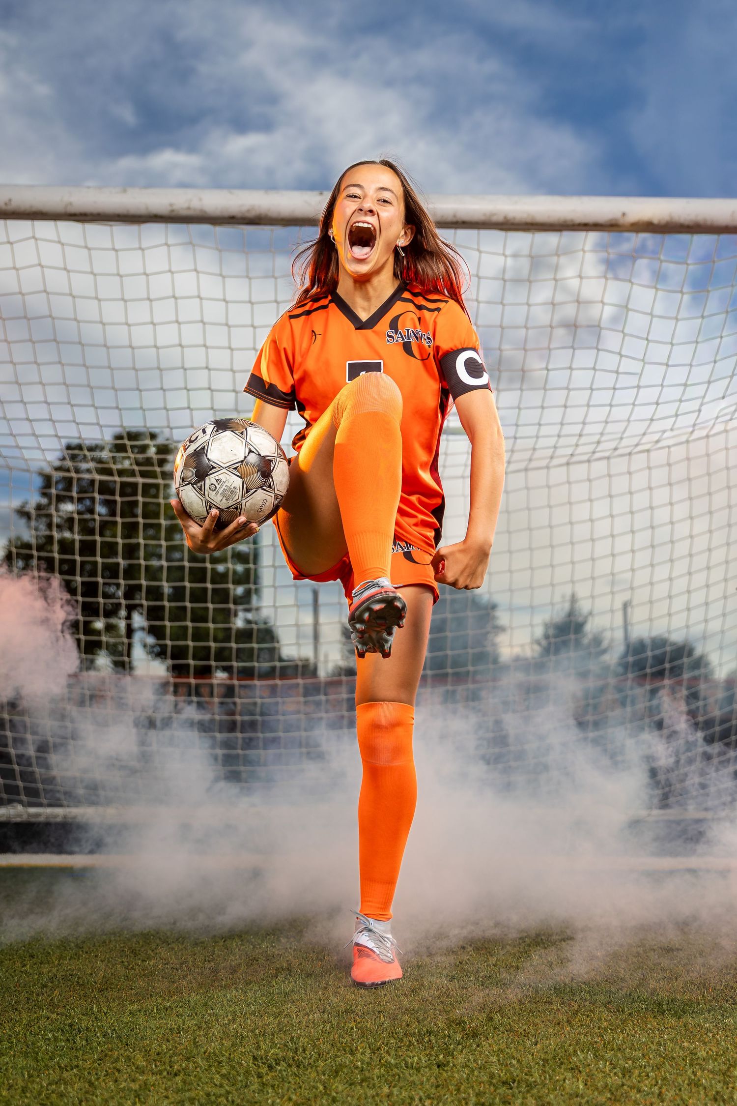Female soccer player in orange uniform kicks ball in front of goal with dramatic smoke effects.