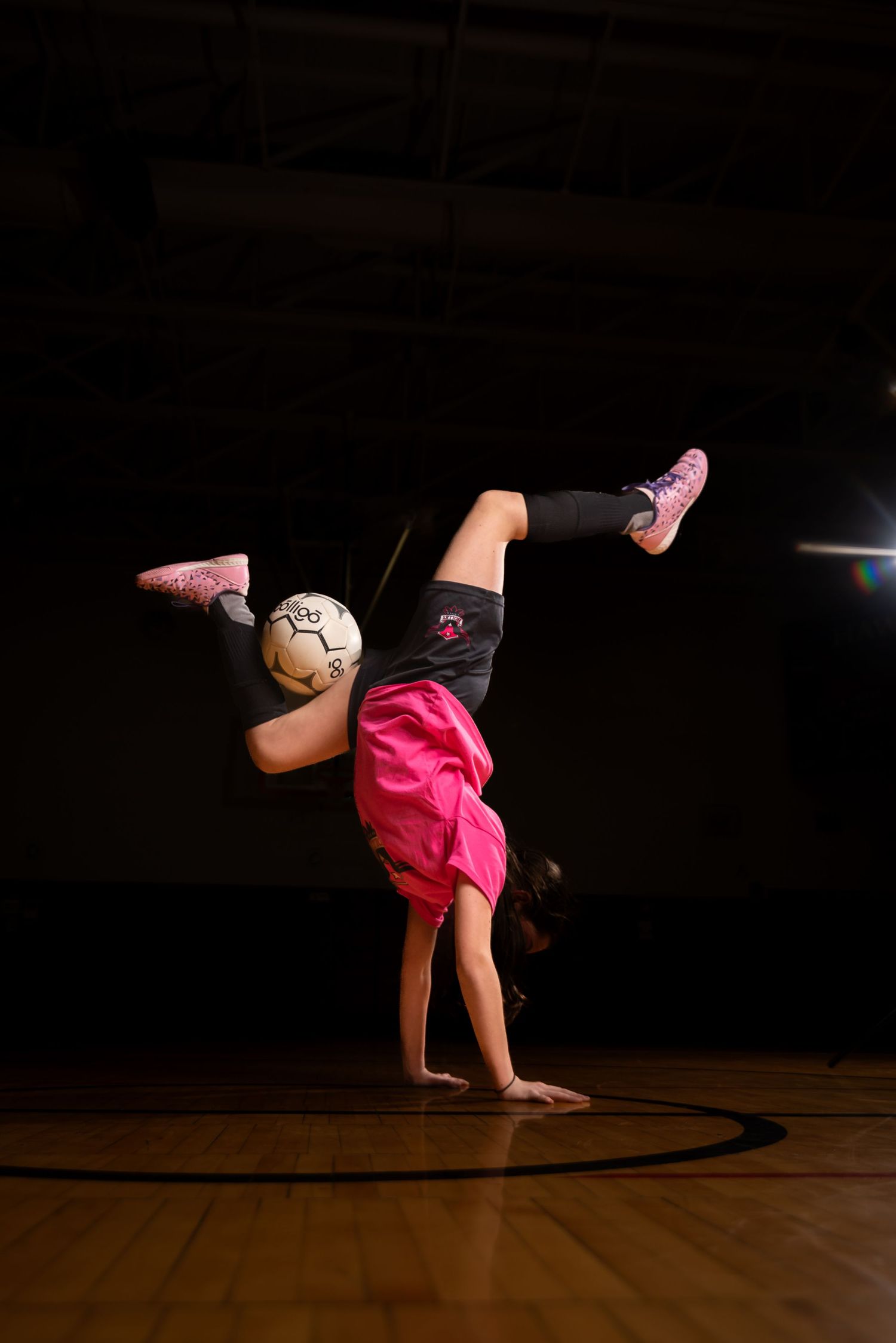 Young girl doing a handstand with a soccer ball balanced on her legs on an indoor court.