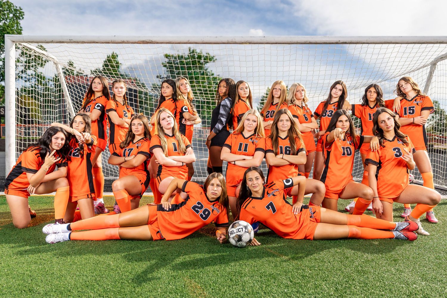 Girls soccer team in orange uniforms posing together in front of a goal net on a green field.