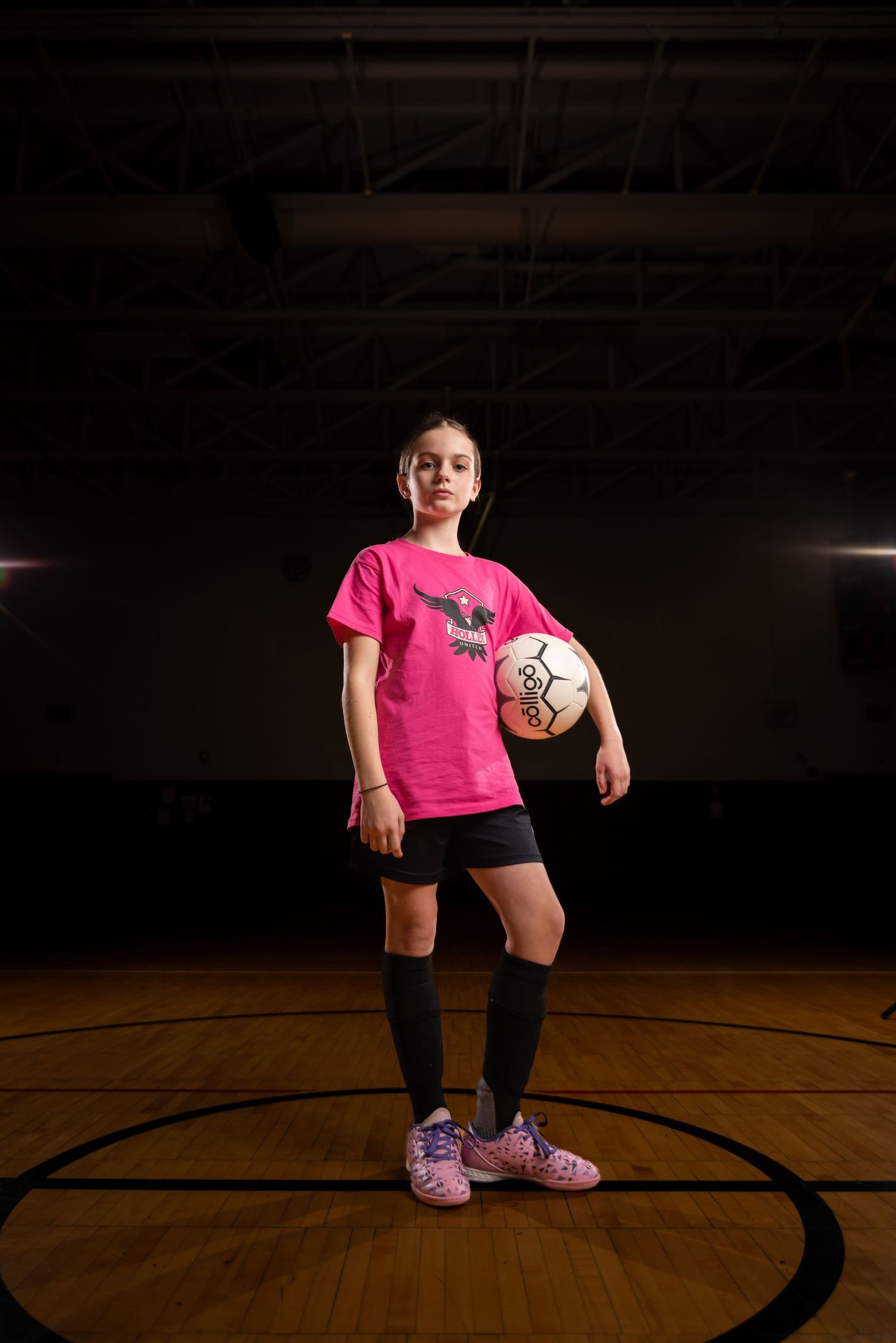 Young girl in pink soccer jersey holds a ball on an indoor court, wearing black shorts and knee-high socks.