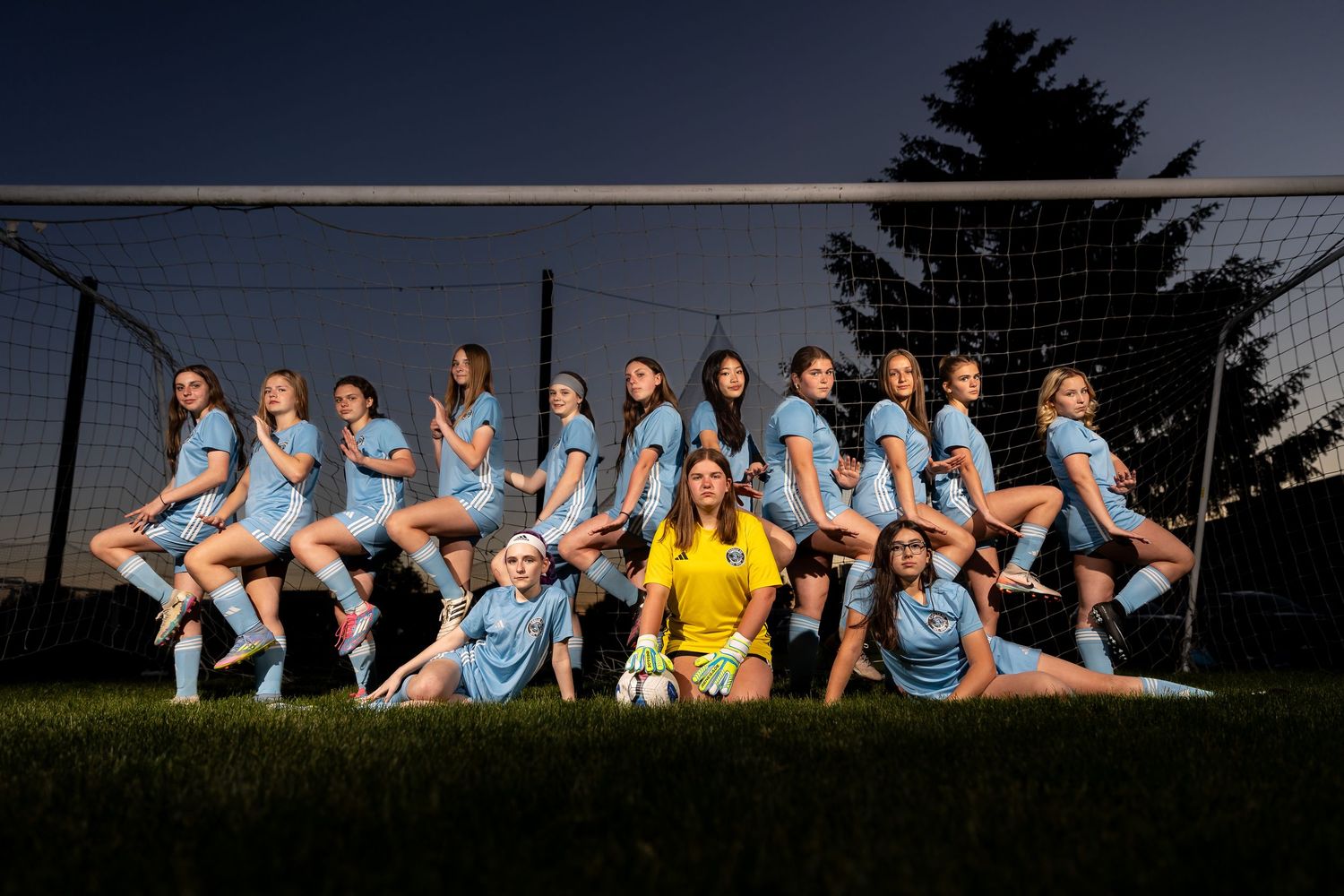 Girls soccer team poses confidently in light blue uniforms in front of a goal net at dusk.