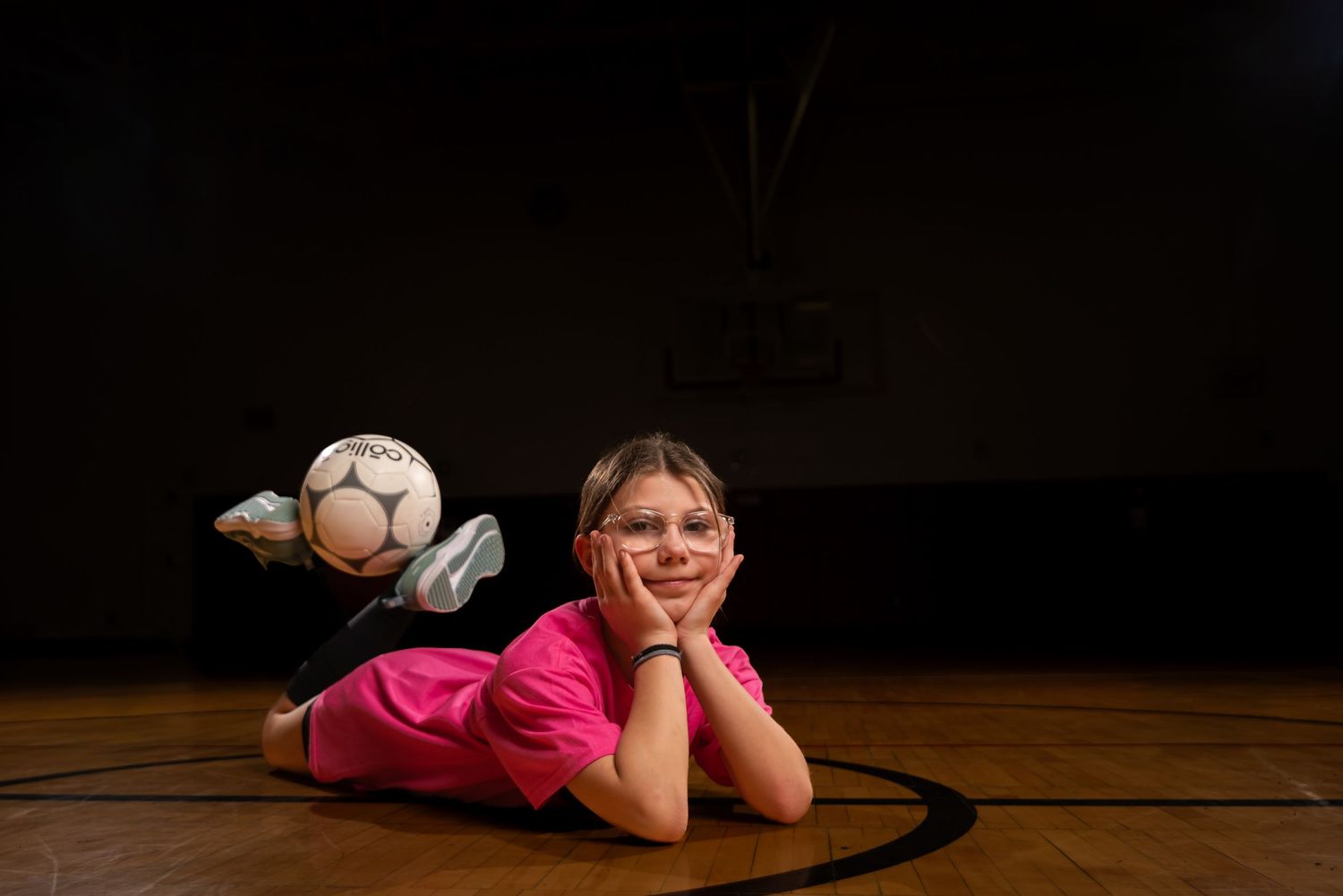 Young girl in pink shirt lying on gym floor with soccer ball, wearing glasses, resting chin on hands.