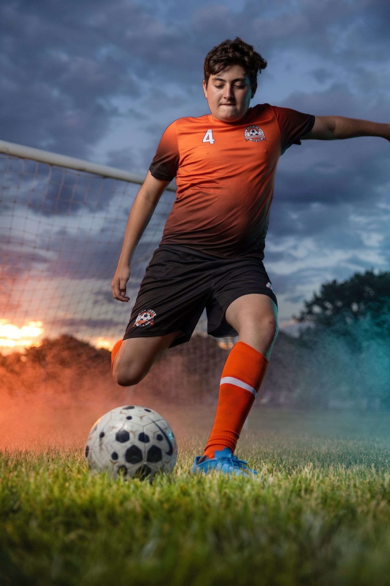 Young soccer player in orange jersey number 4 kicking a ball on field at dusk with dramatic colorful smoke.