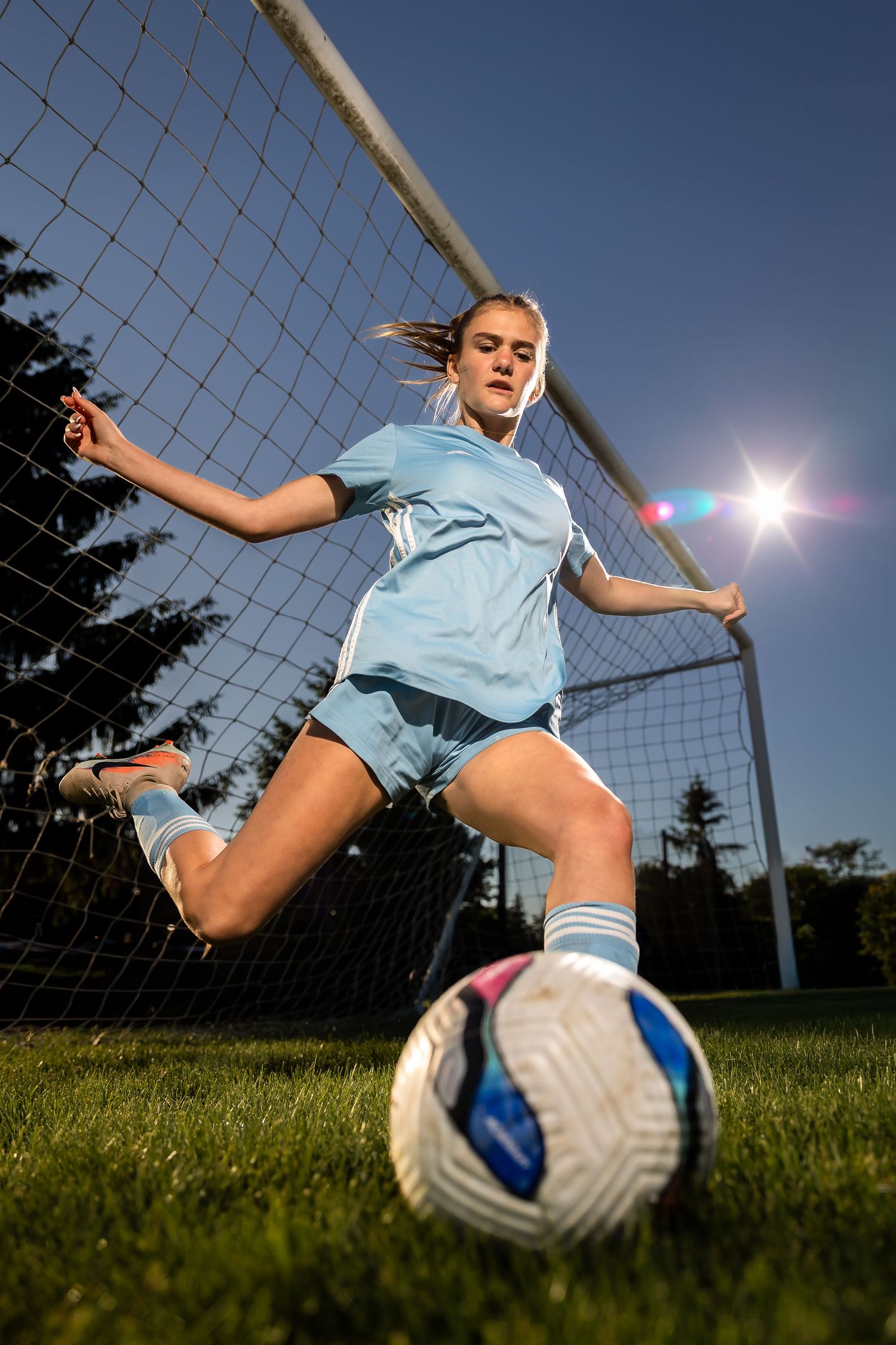 Young female soccer player in light blue uniform about to kick a soccer ball in front of a goal net outdoors.