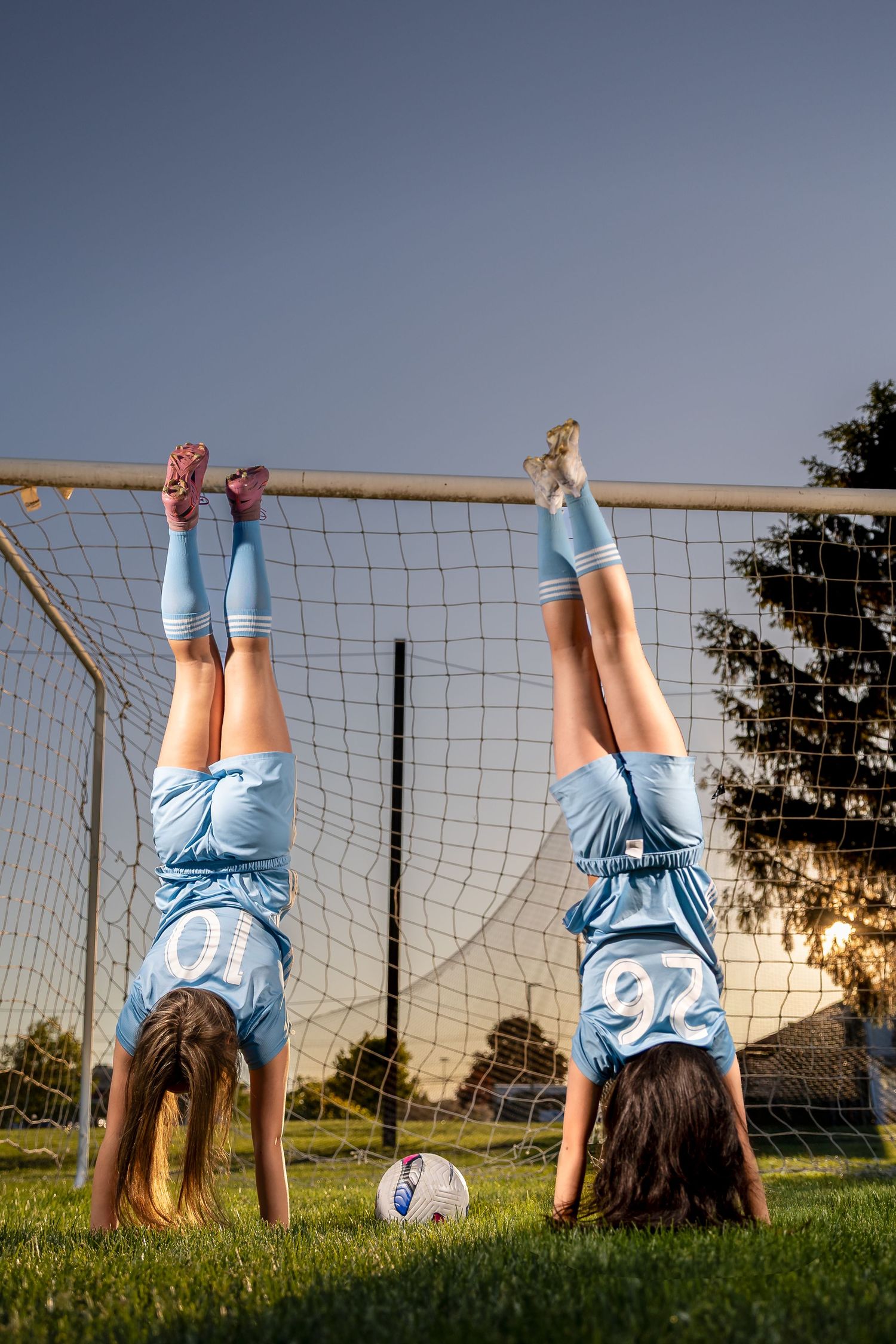 Two girls in blue soccer uniforms doing handstands on a goal post with a soccer ball on the grass.