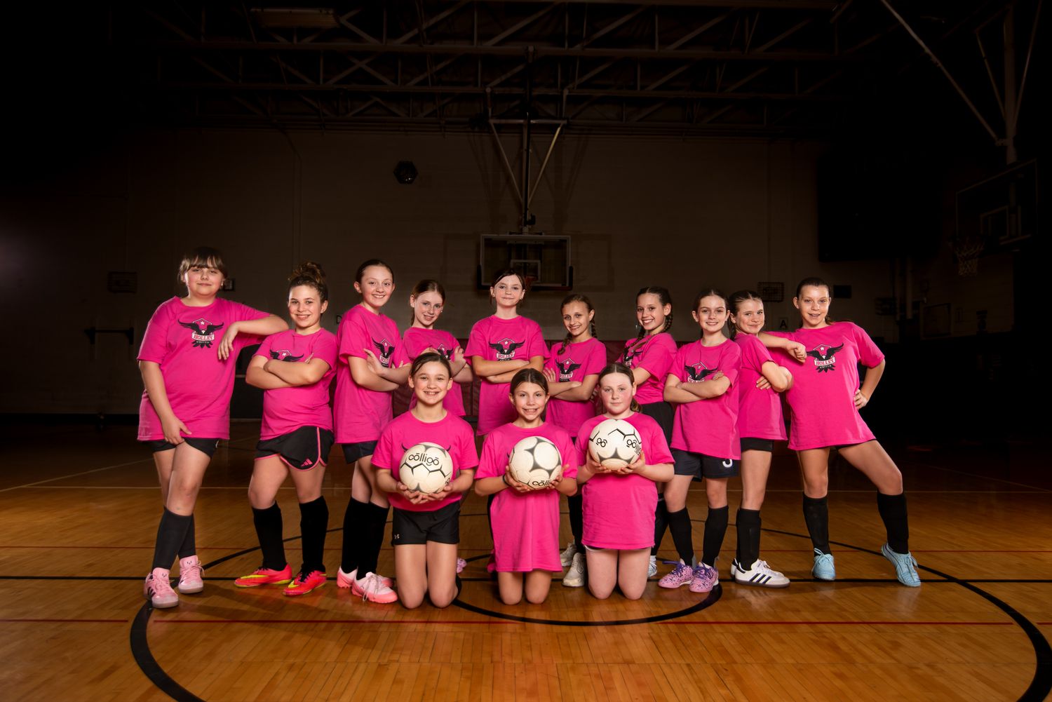 Girls youth soccer team in pink jerseys posing with soccer balls on an indoor basketball court.