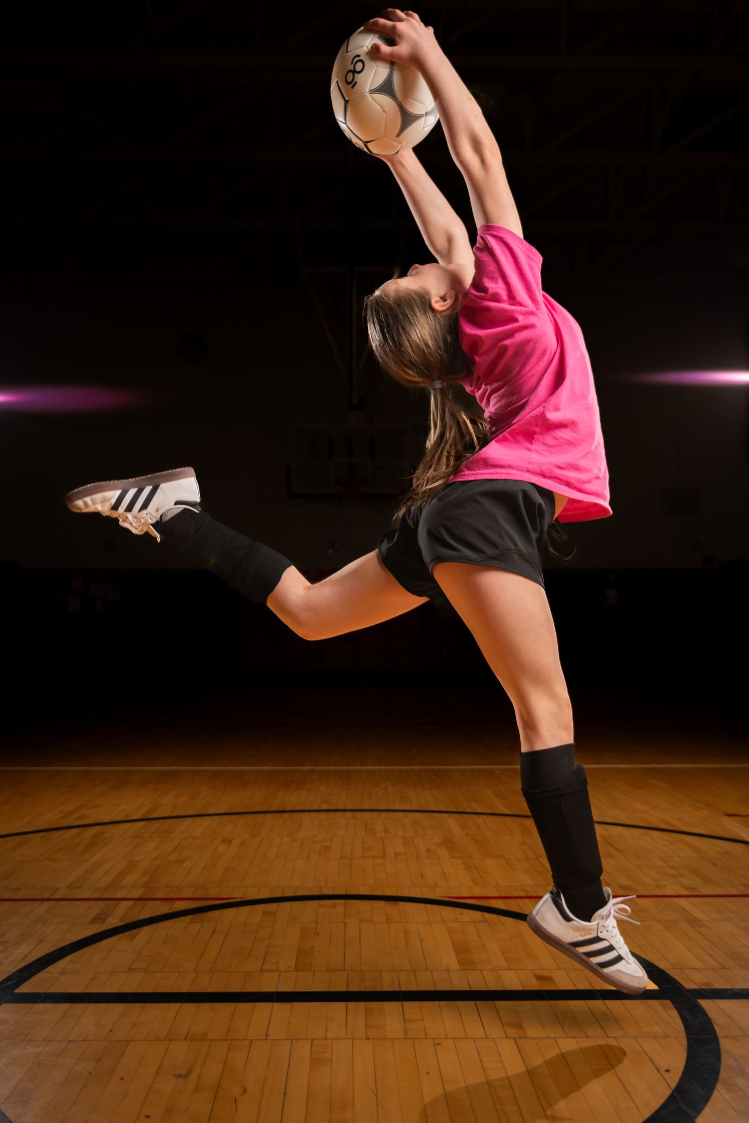 Female volleyball player in pink jersey leaping to spike a ball on an indoor hardwood court.