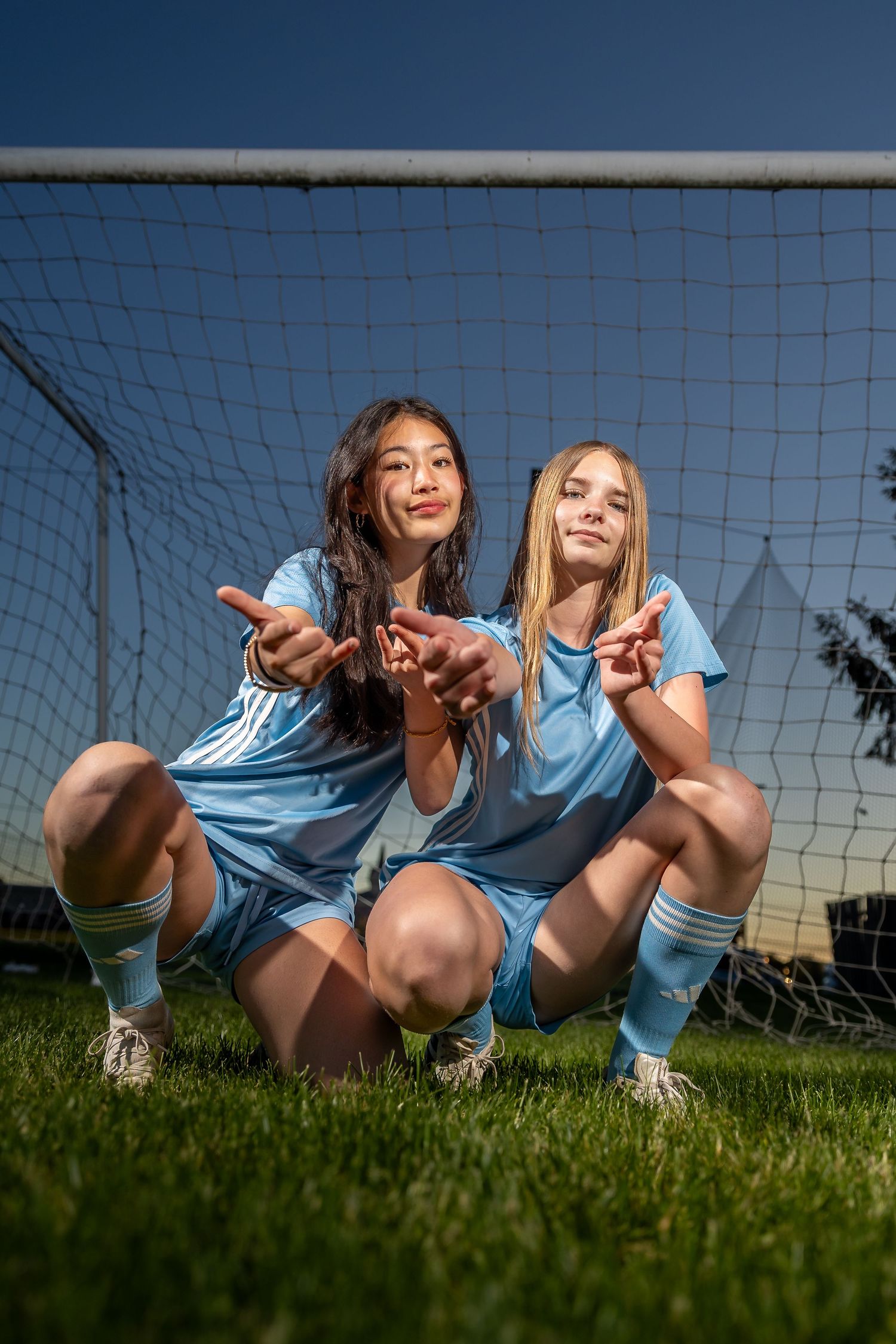 Two girls in blue soccer uniforms posing in front of a goal net on a grass field.