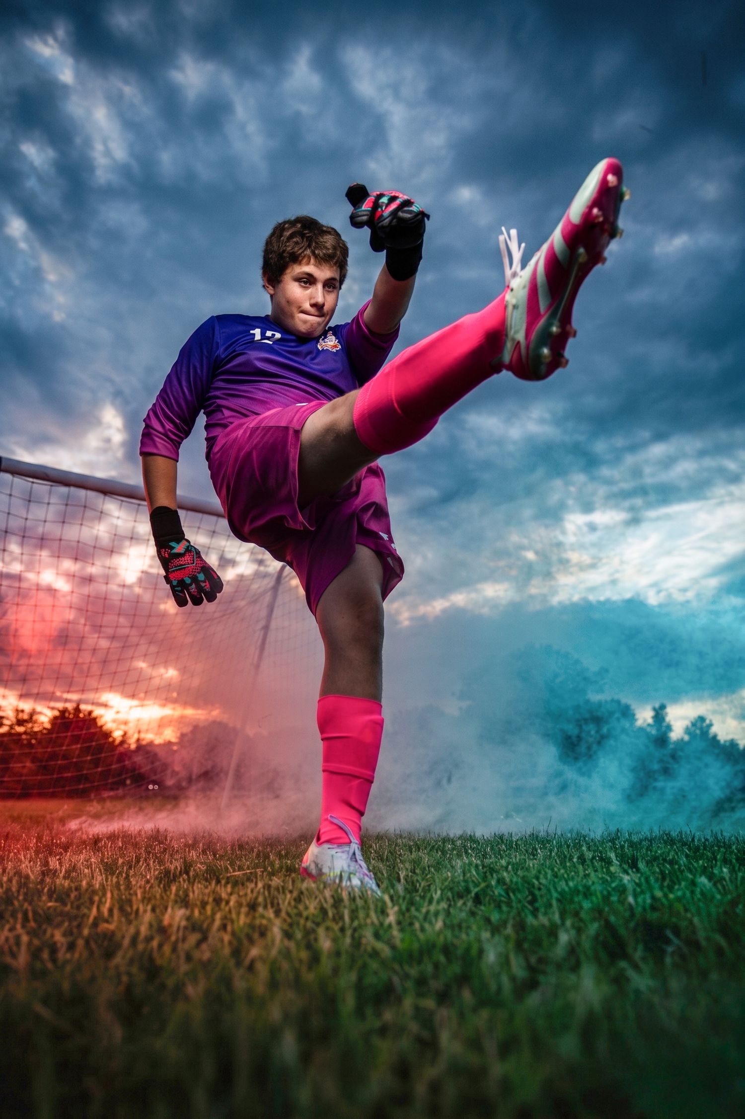 Soccer goalkeeper in purple and pink uniform kicking ball on field with colorful smoke and dramatic sky.