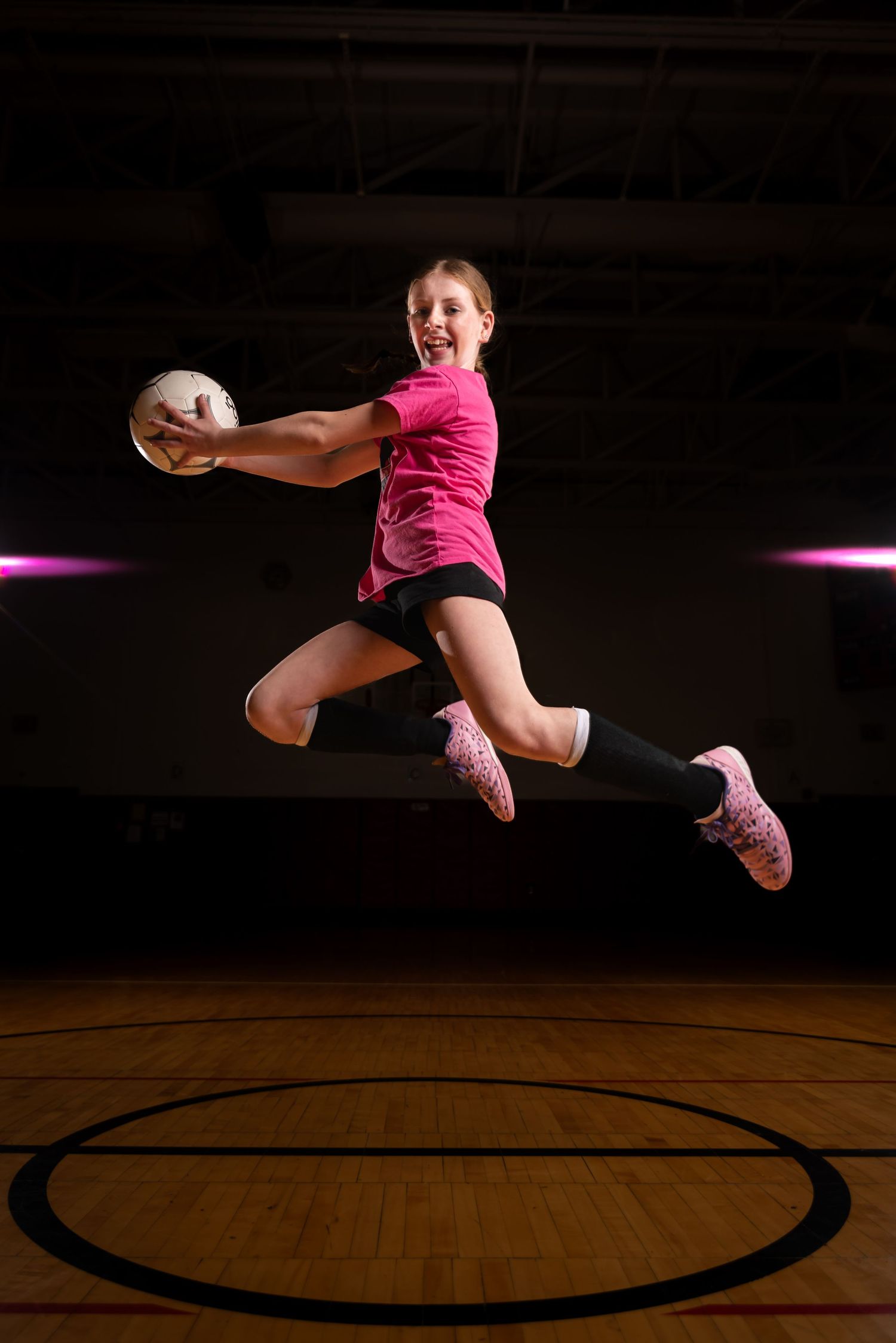 Young girl in pink jersey leaps mid-air holding volleyball in dark gymnasium, wearing pink shoes and knee socks.