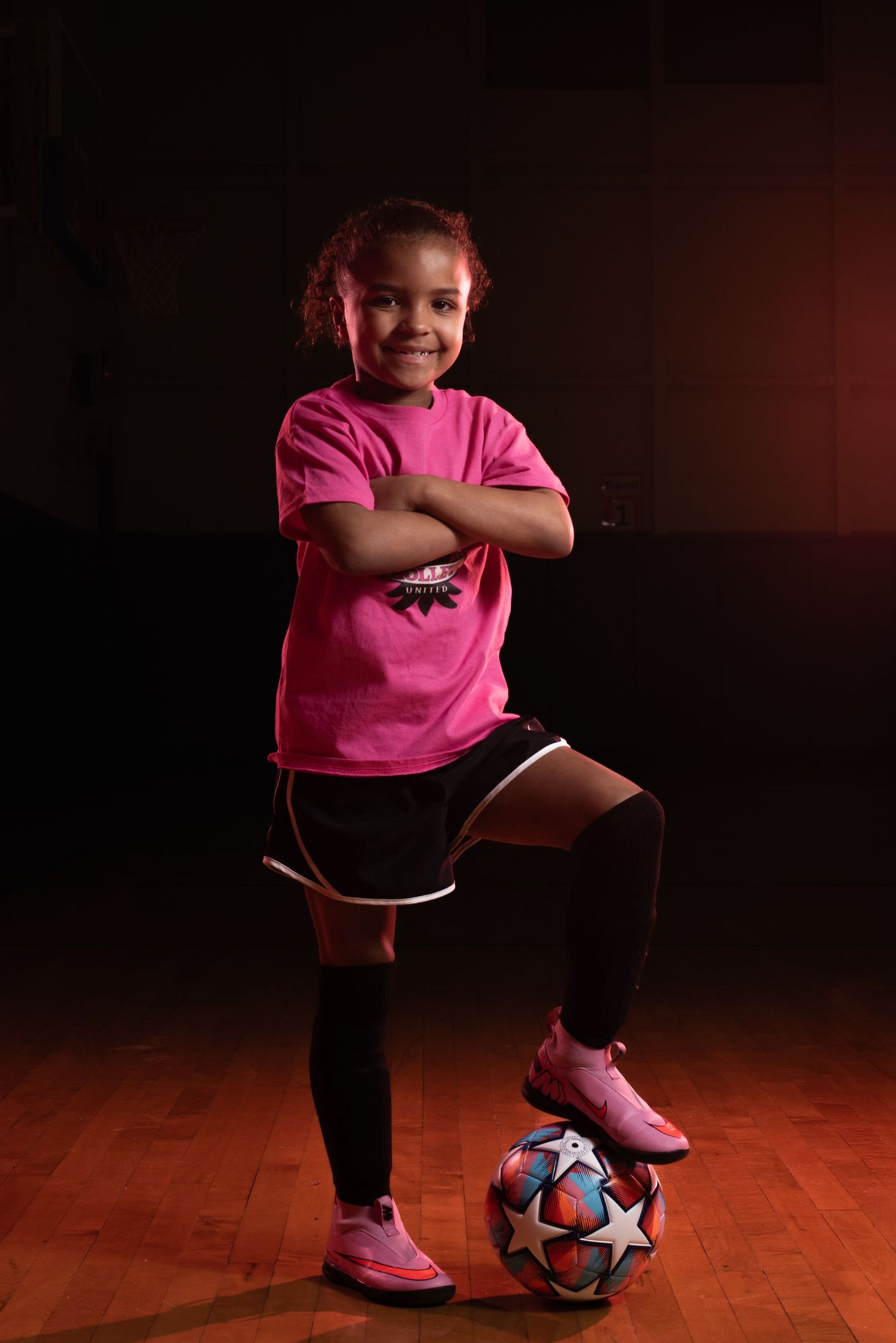 Soccer player in pink shirt poses confidently with ball in dramatic studio lighting. Studio portrait of a youth athlete. Team photos.