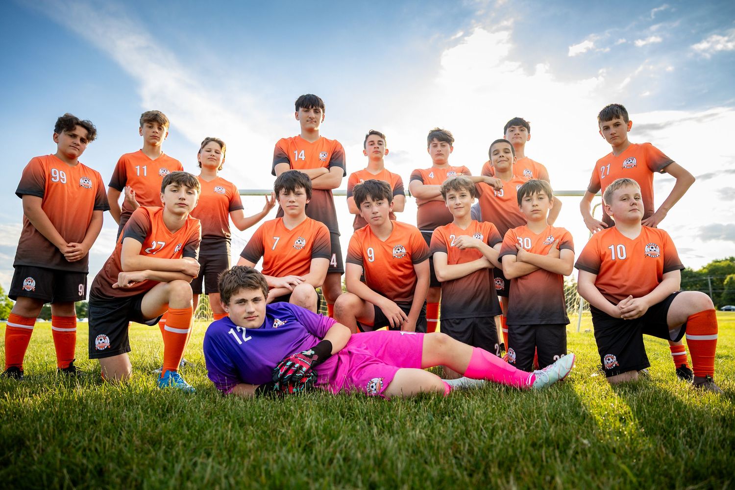 Youth soccer team poses on grass field, wearing orange uniforms, with goalkeeper in purple and pink lying in front.