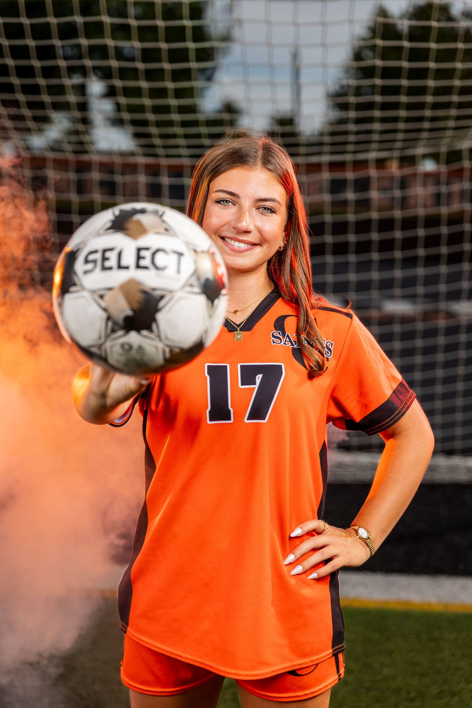 A smiling female soccer player in an orange #17 jersey holds a Select ball toward the camera with colorful smoke in the background.
