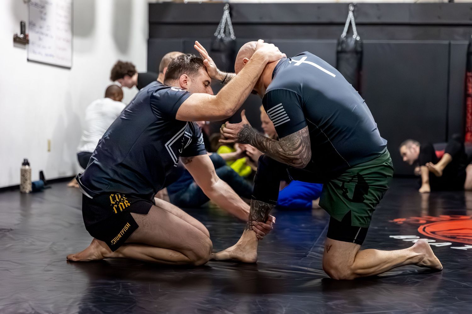 Two men practicing grappling techniques on a black mat in a martial arts gym during training.
