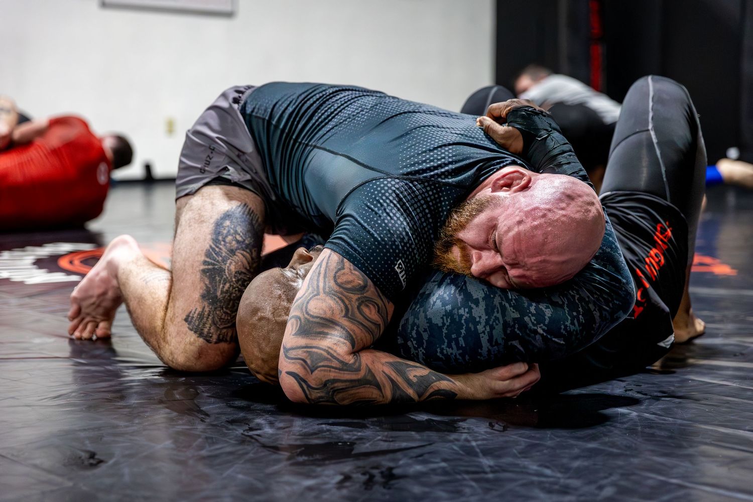 Two grapplers practice Brazilian jiu-jitsu ground techniques on a black mat in a martial arts gym.