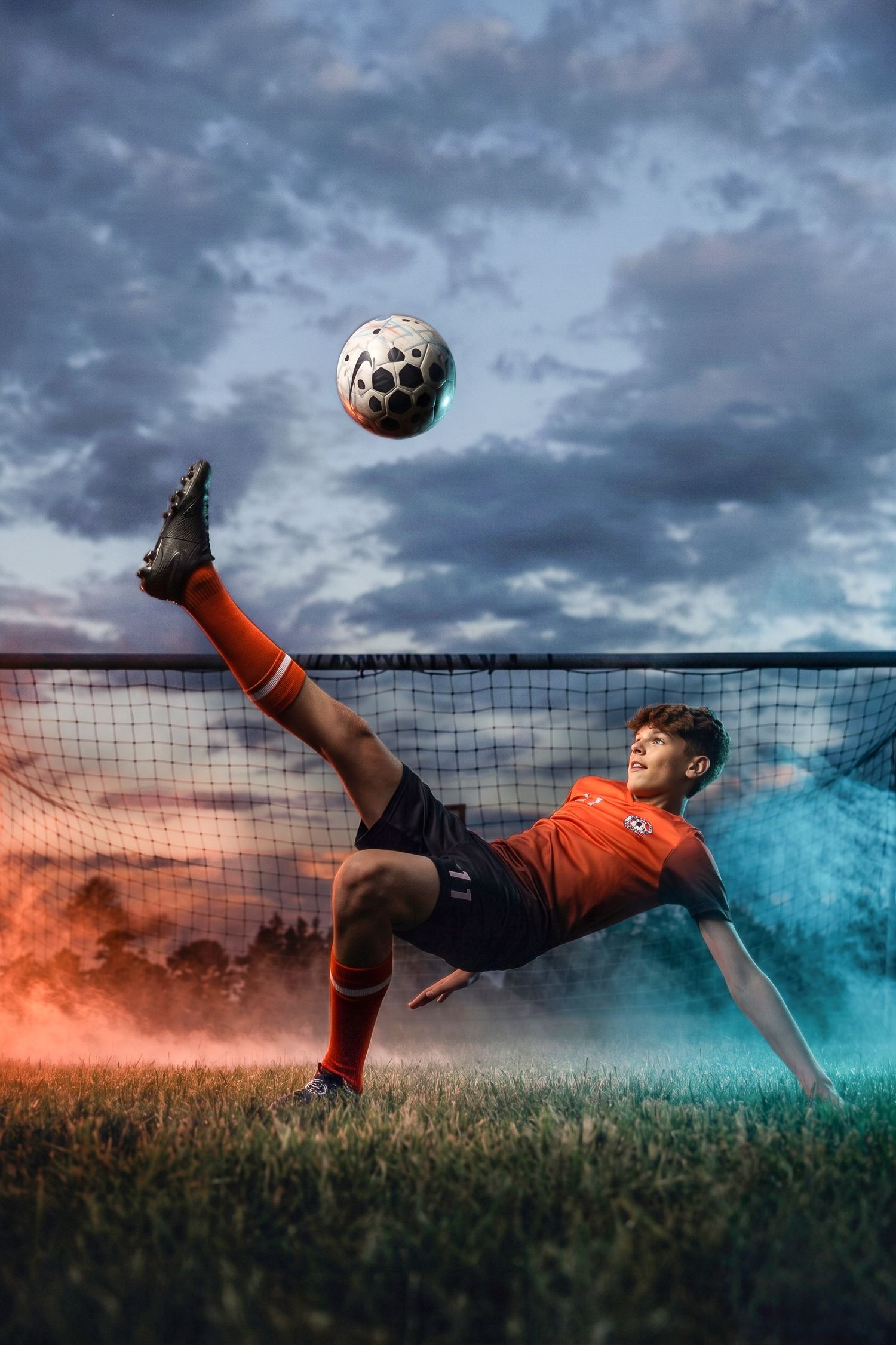 Soccer player in orange jersey performs a bicycle kick in front of a goal with dramatic cloudy sky and colorful smoke.