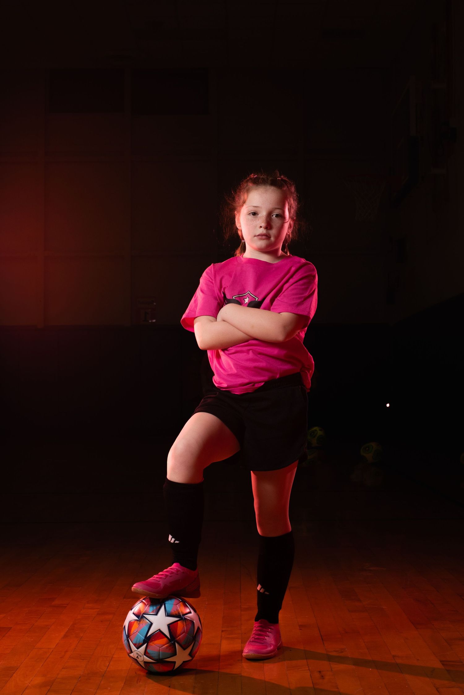 Soccer player in pink shirt and black shorts poses with ball against dark background. Studio portrait of a youth athlete. Team photos.