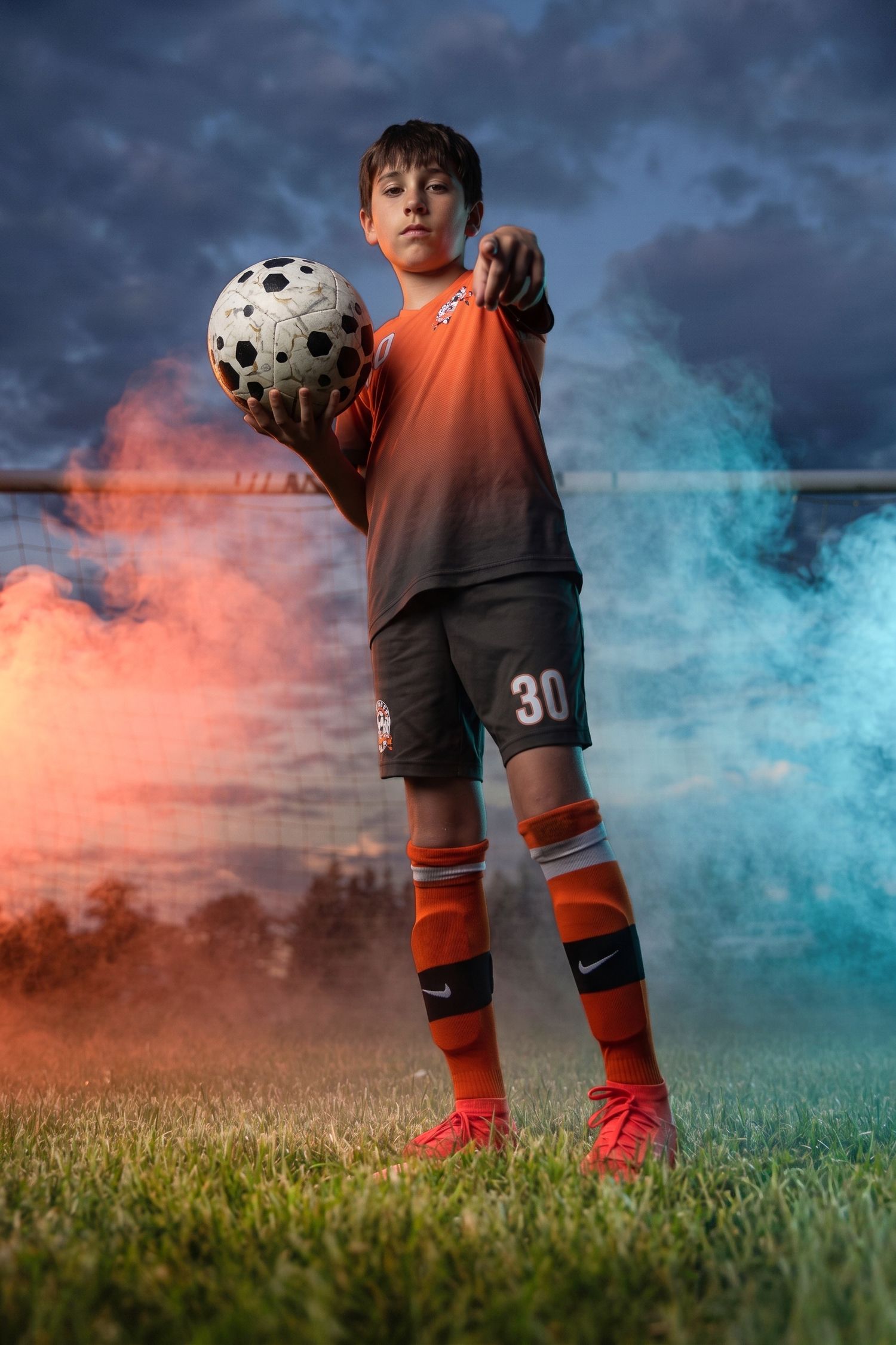 Boy in orange soccer uniform #30 holds ball and points at camera on field with colorful smoke at dusk.