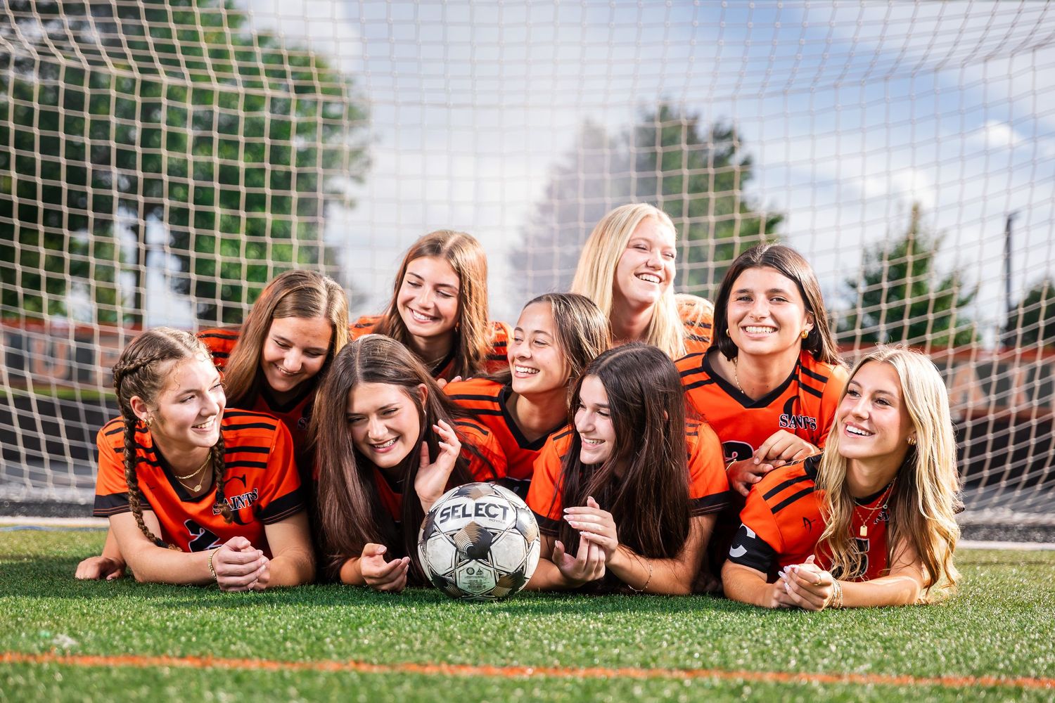 Girls soccer team posing together in front of a goal net on a green field, wearing orange and black uniforms.