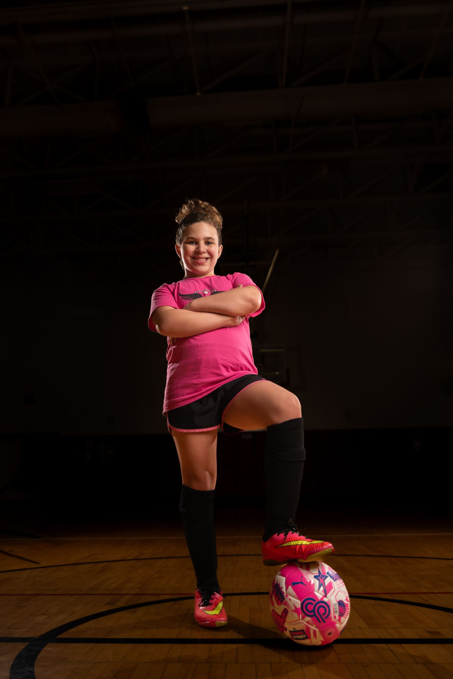 Young female soccer player in pink jersey poses confidently with foot on ball in indoor gym.