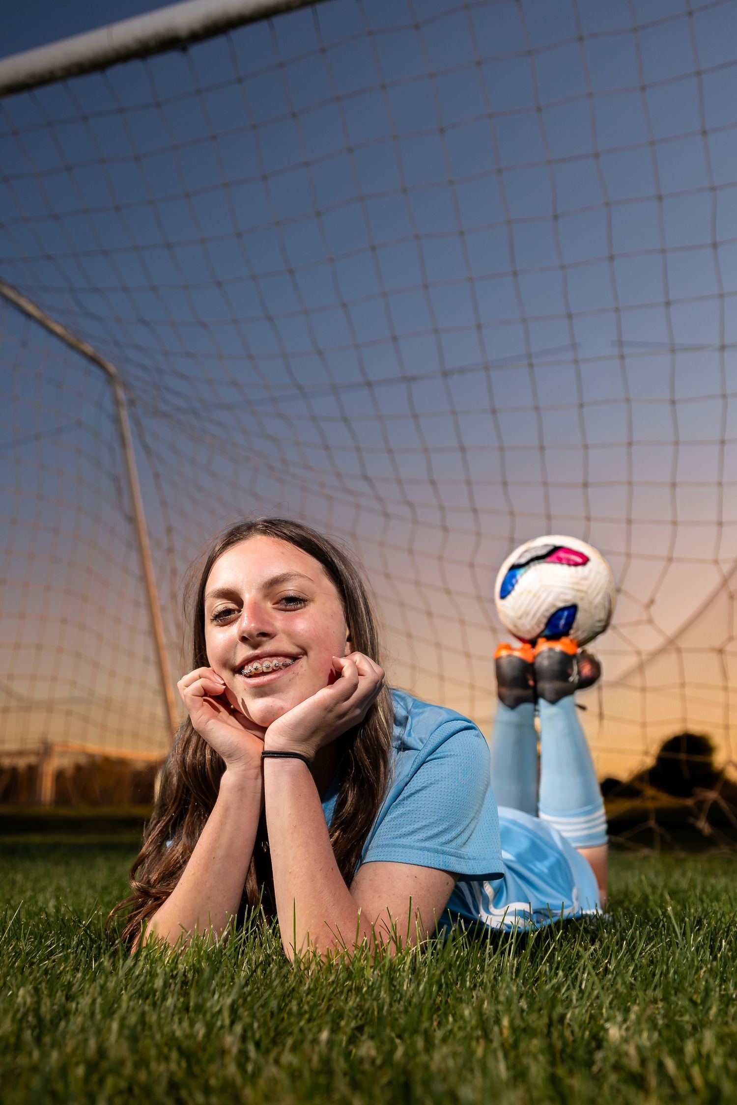 Young female soccer player lying on grass field, smiling in blue uniform, holding soccer ball near goal net at sunset.